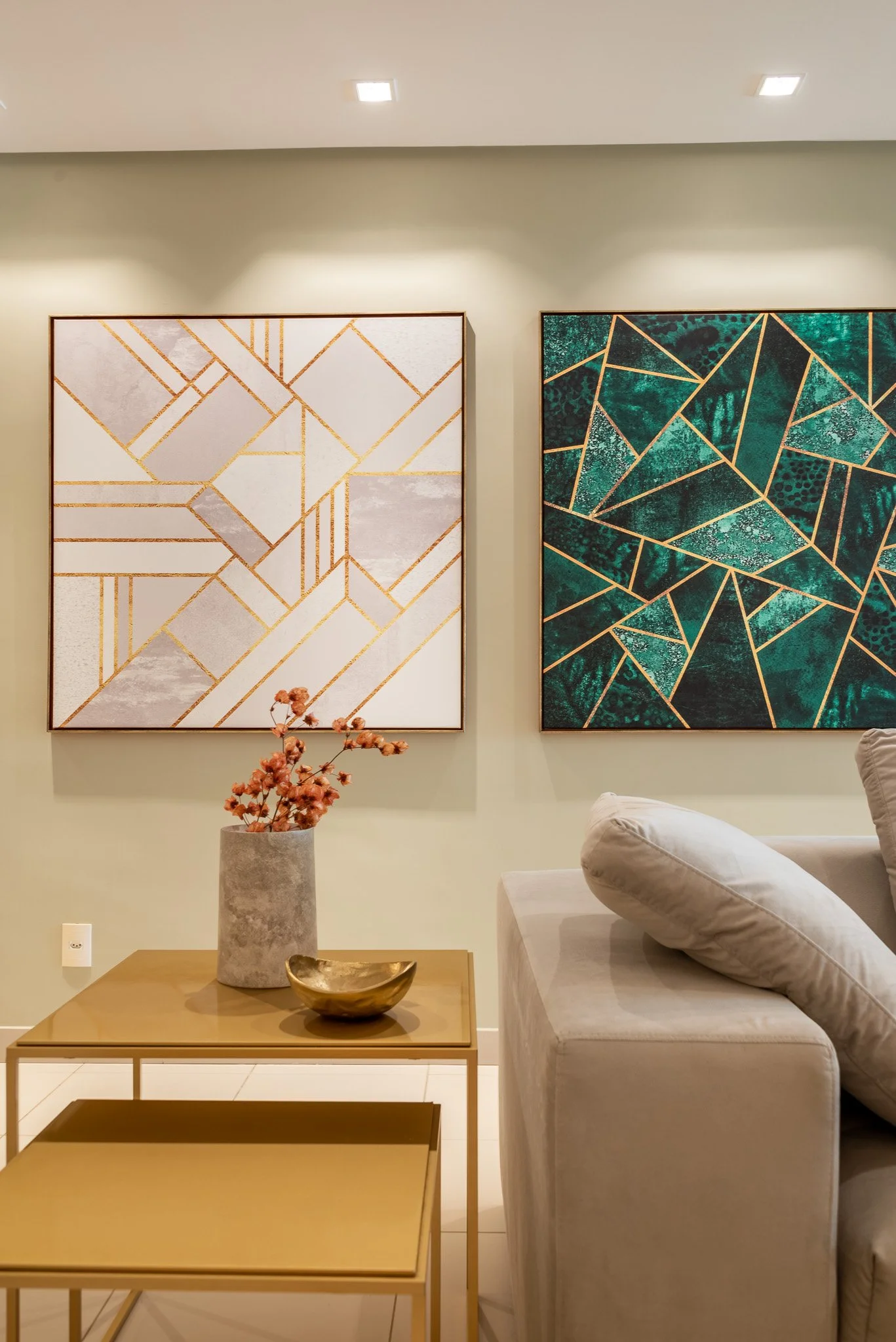 Living room with two modern geometric art pieces on the wall, a beige sofa, a wooden coffee table with a vase of pink flowers, and a gold decorative bowl.