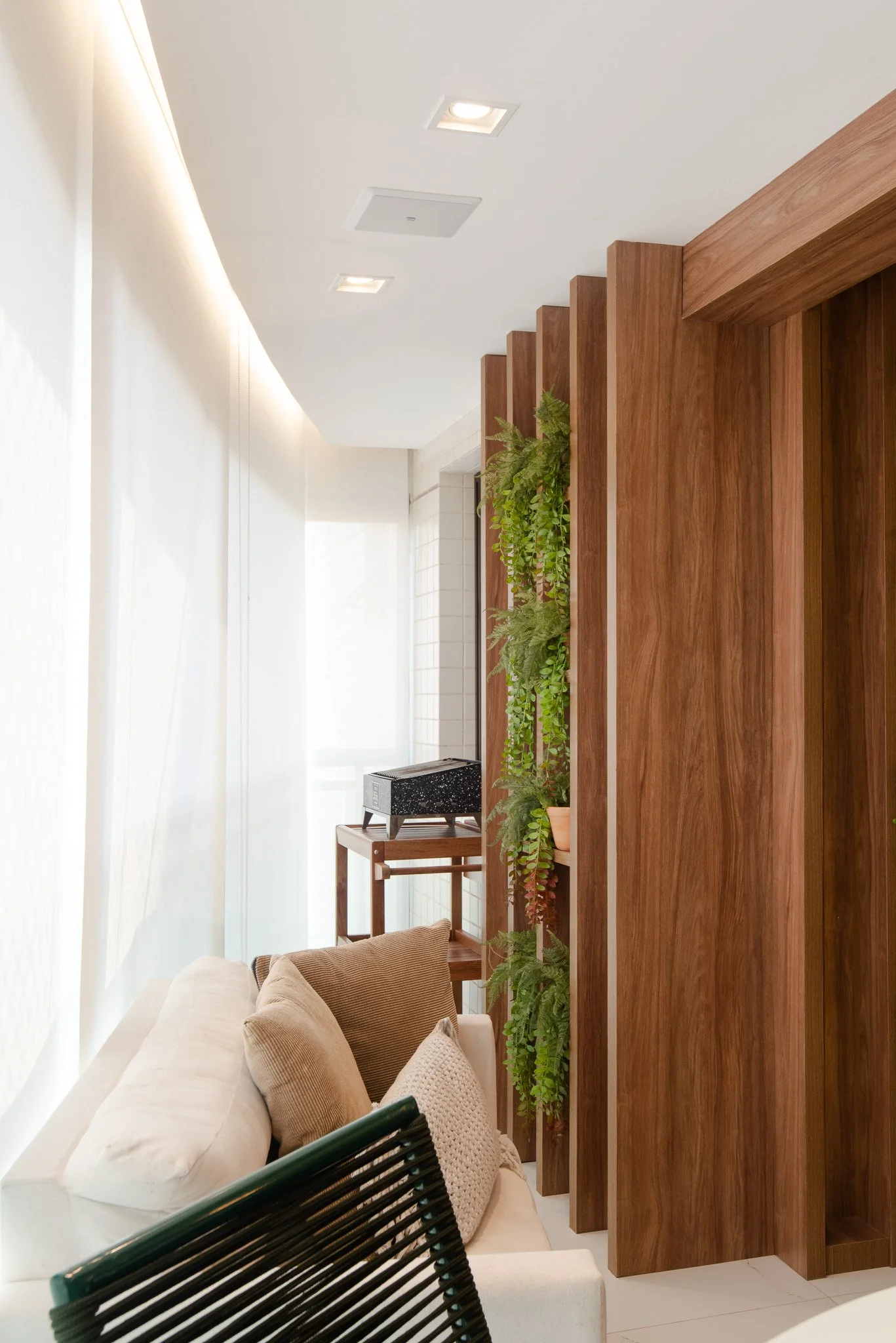 Living room corner with beige couch, cushions, wooden wall with green plants, window with sheer curtains, ceiling lights, and a small table with a black box.