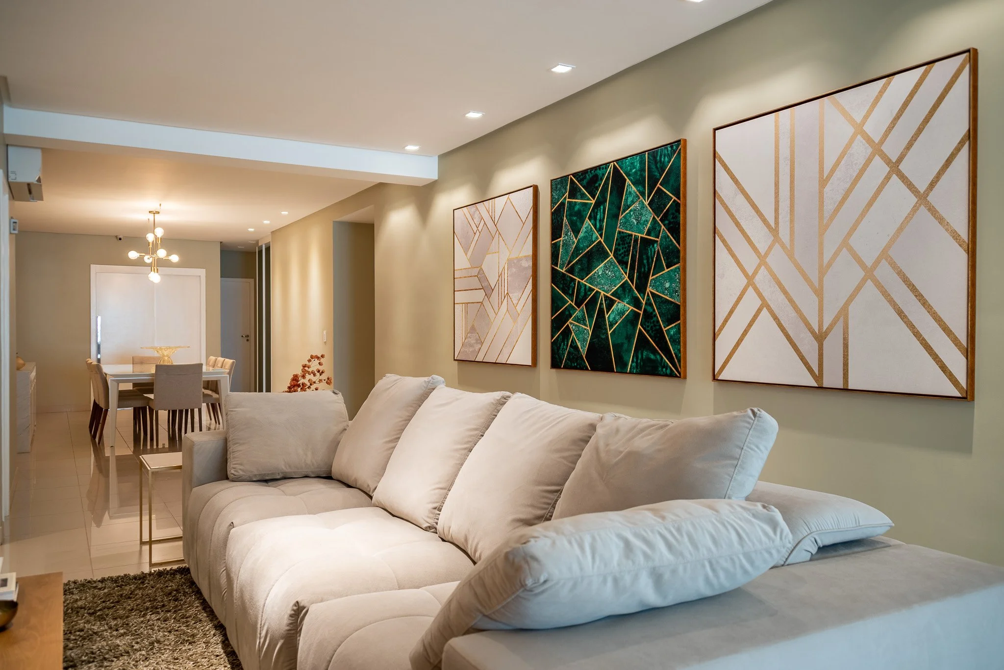 Living room with beige sofa, three abstract art paintings with gold accents on the wall, and a dining area in the background with modern chandelier.