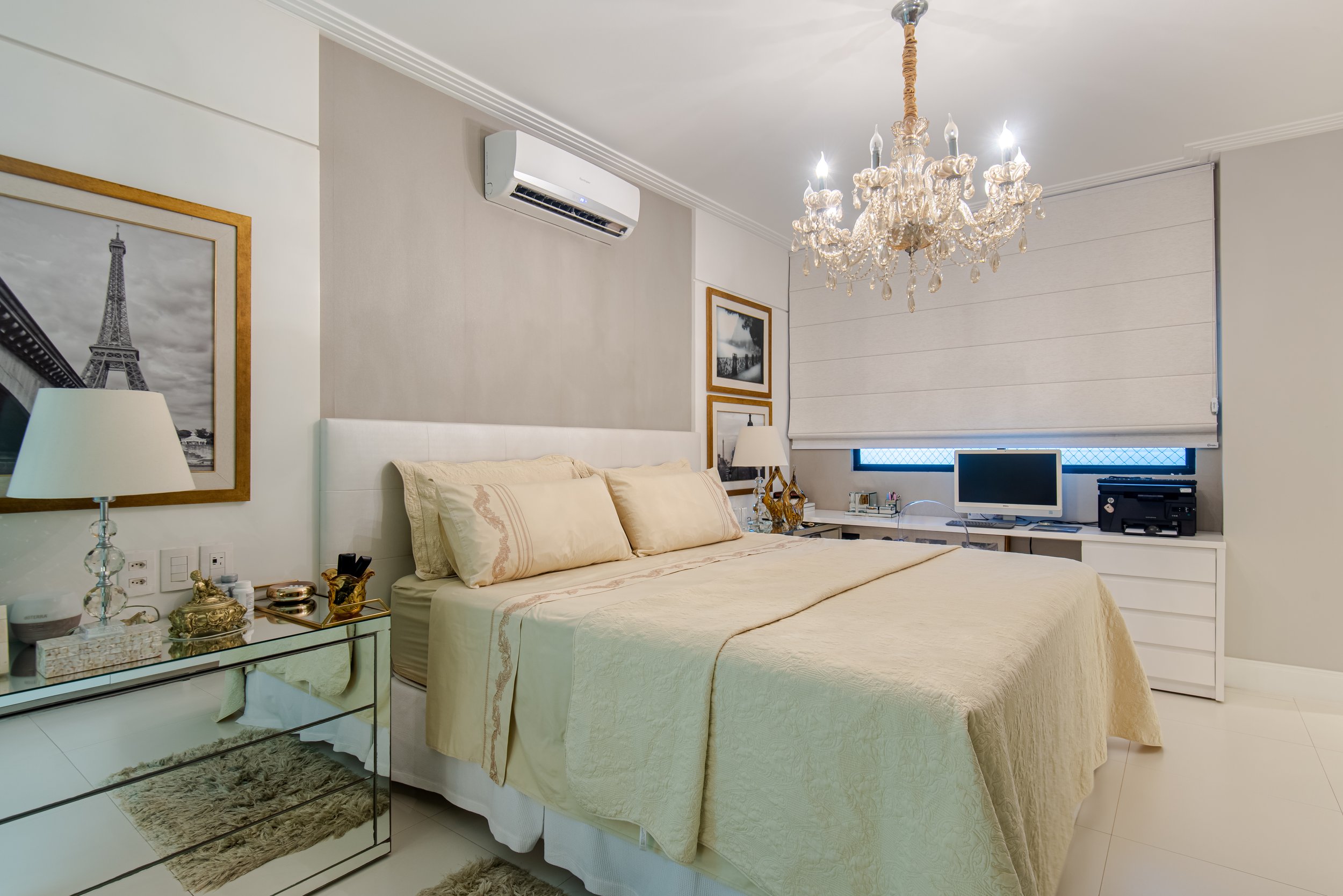 Elegant bedroom with cream bedding, white headboard, mirrored side table, framed black and white Paris Eiffel Tower picture, chandelier, air conditioning unit, window with blinds, desk with computer and printer, and modern decor.