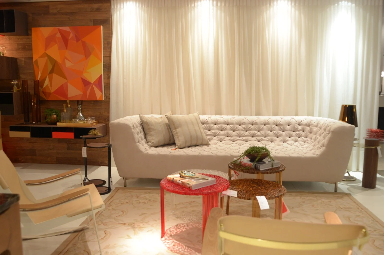 A living room with a large white tufted sofa with four pillows, a beige rug, and three small tables with various decorations. There is a armchair to the left and a wooden accent wall with a colorful geometric artwork. The room has sheer white curtain