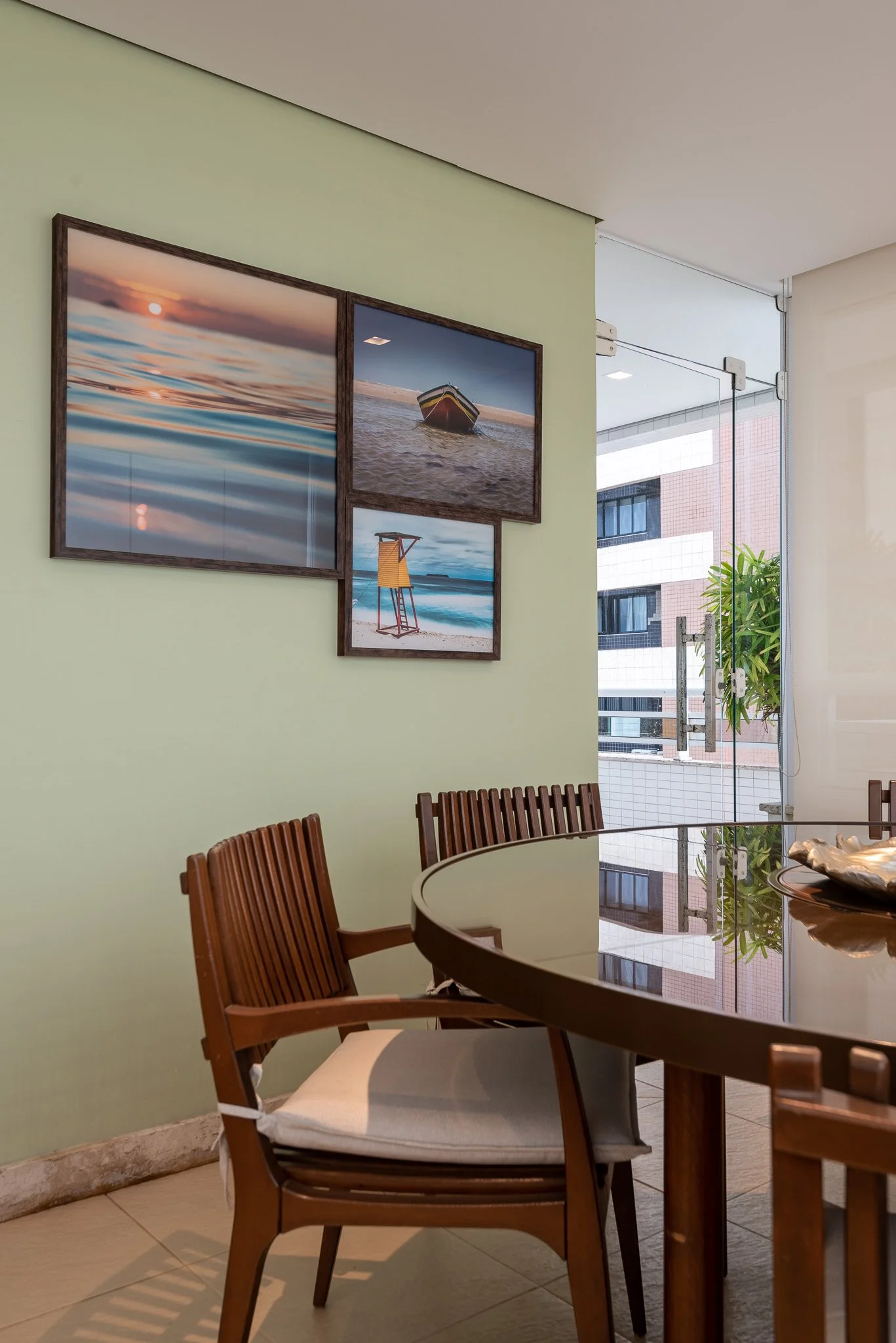 Inside a room with a green wall decorated with four framed beach-themed photographs, a round wooden table, and wooden chairs with white cushions, next to a glass door with a city building visible outside.