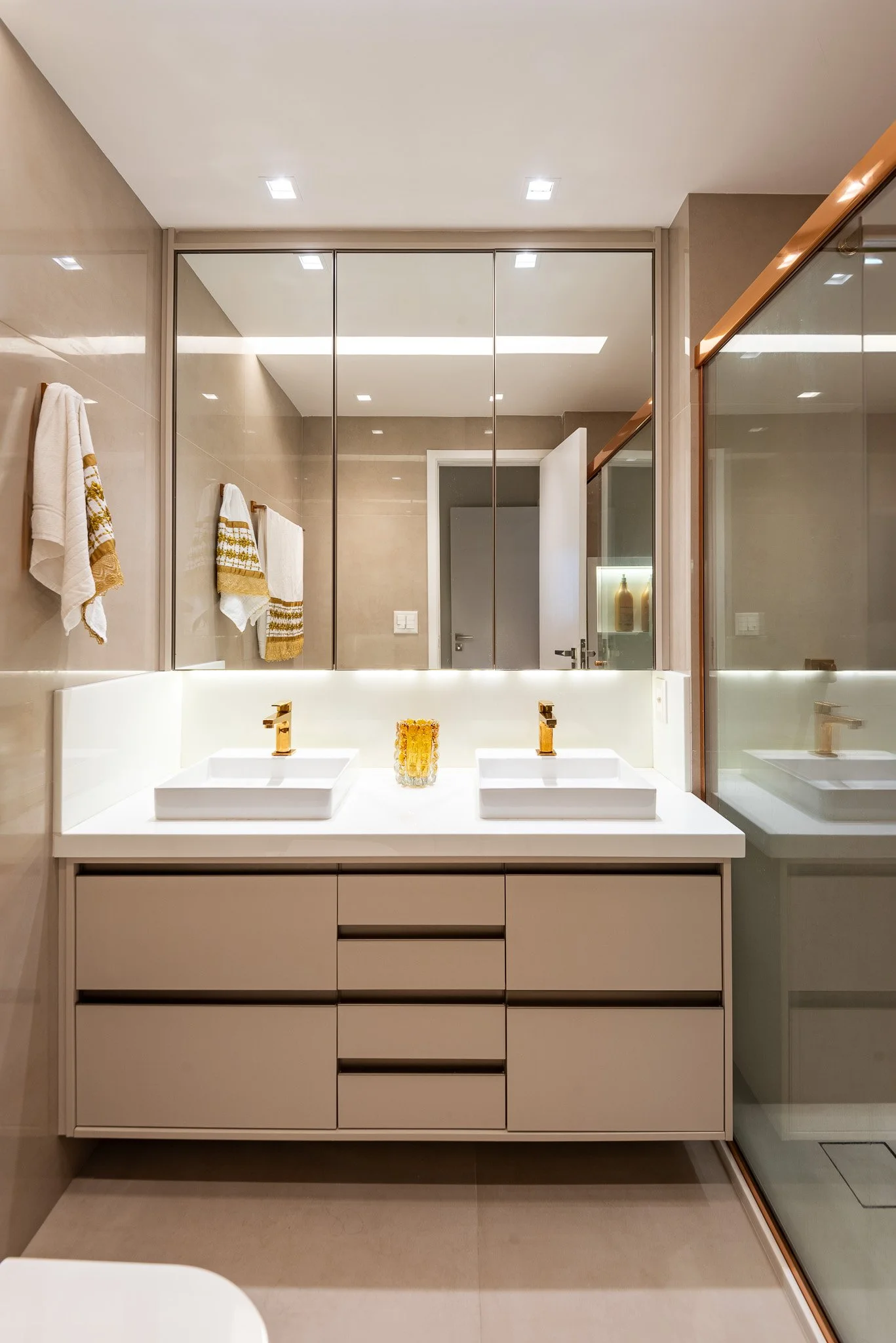 Modern bathroom with double vanity, white sinks, gold fixtures, large mirror, beige tiles, towels on hooks, glass shower with wooden trim.