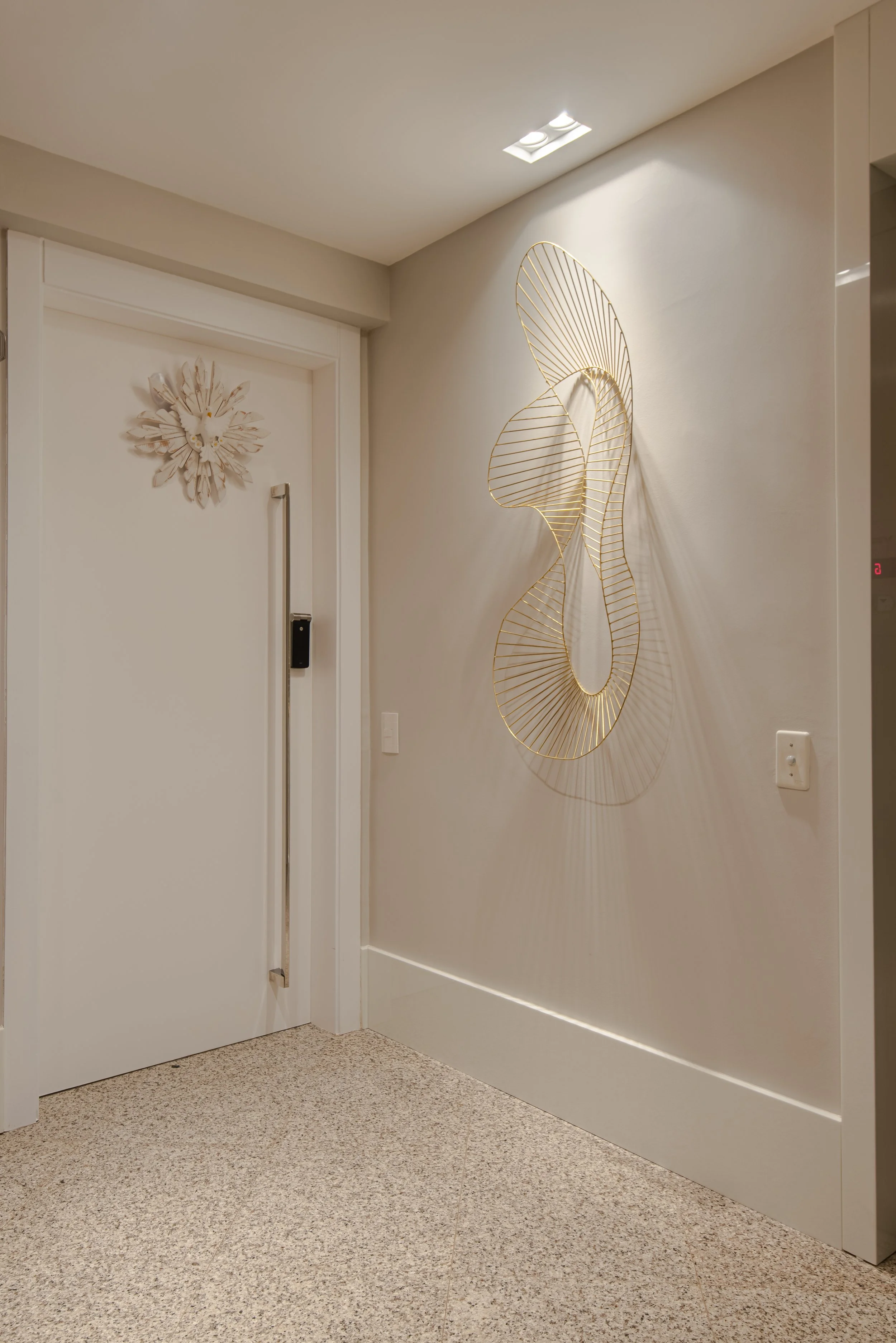 Interior view of a hallway corner with a closed white door with a decorative flower or sunburst wall ornament above the door handle, and a modern metal wall sculpture on the right wall shaped like an abstract figure with shadow cast on the wall. The 