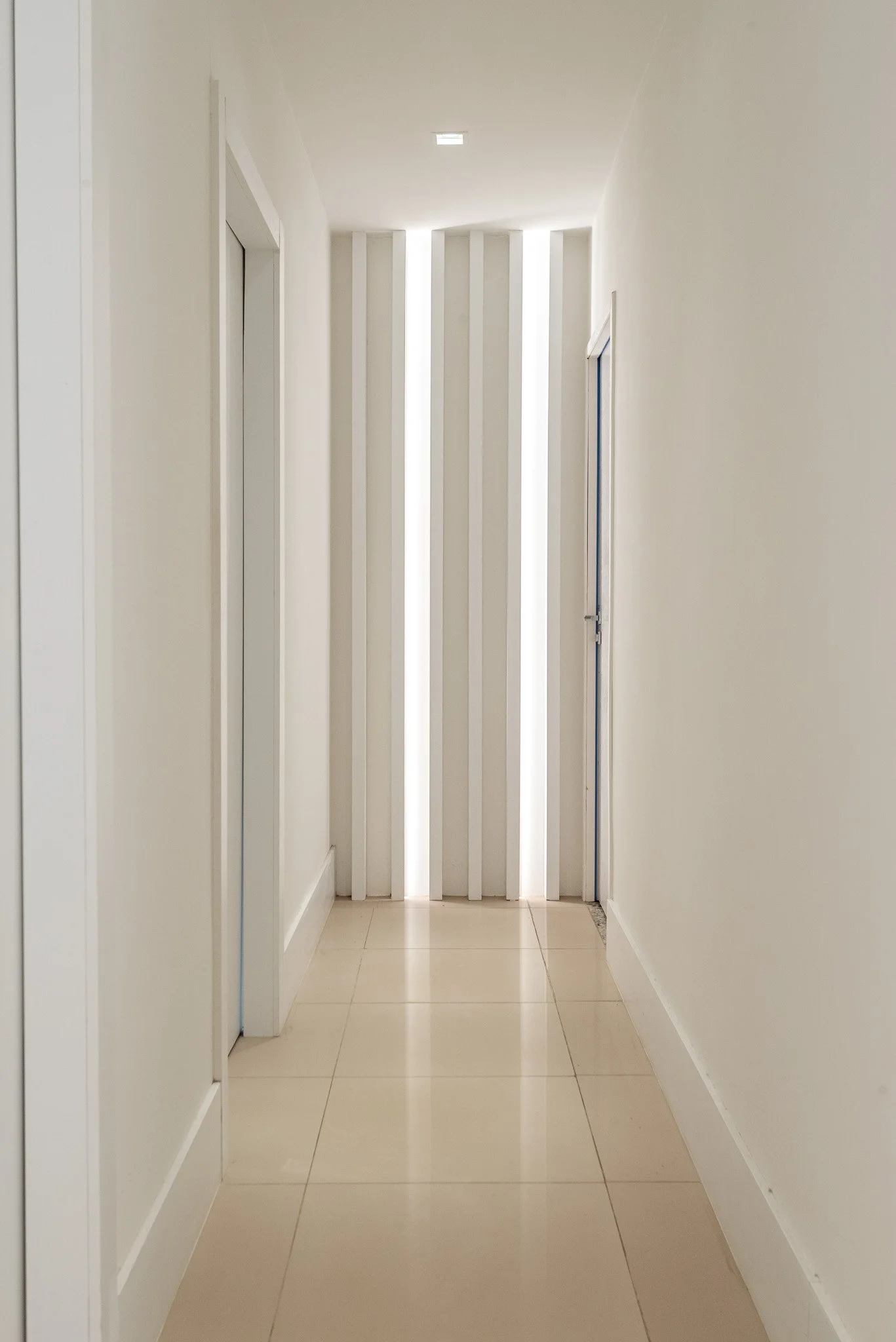Empty hallway with white walls, beige tile floor, and a door at the end with vertical light strips.