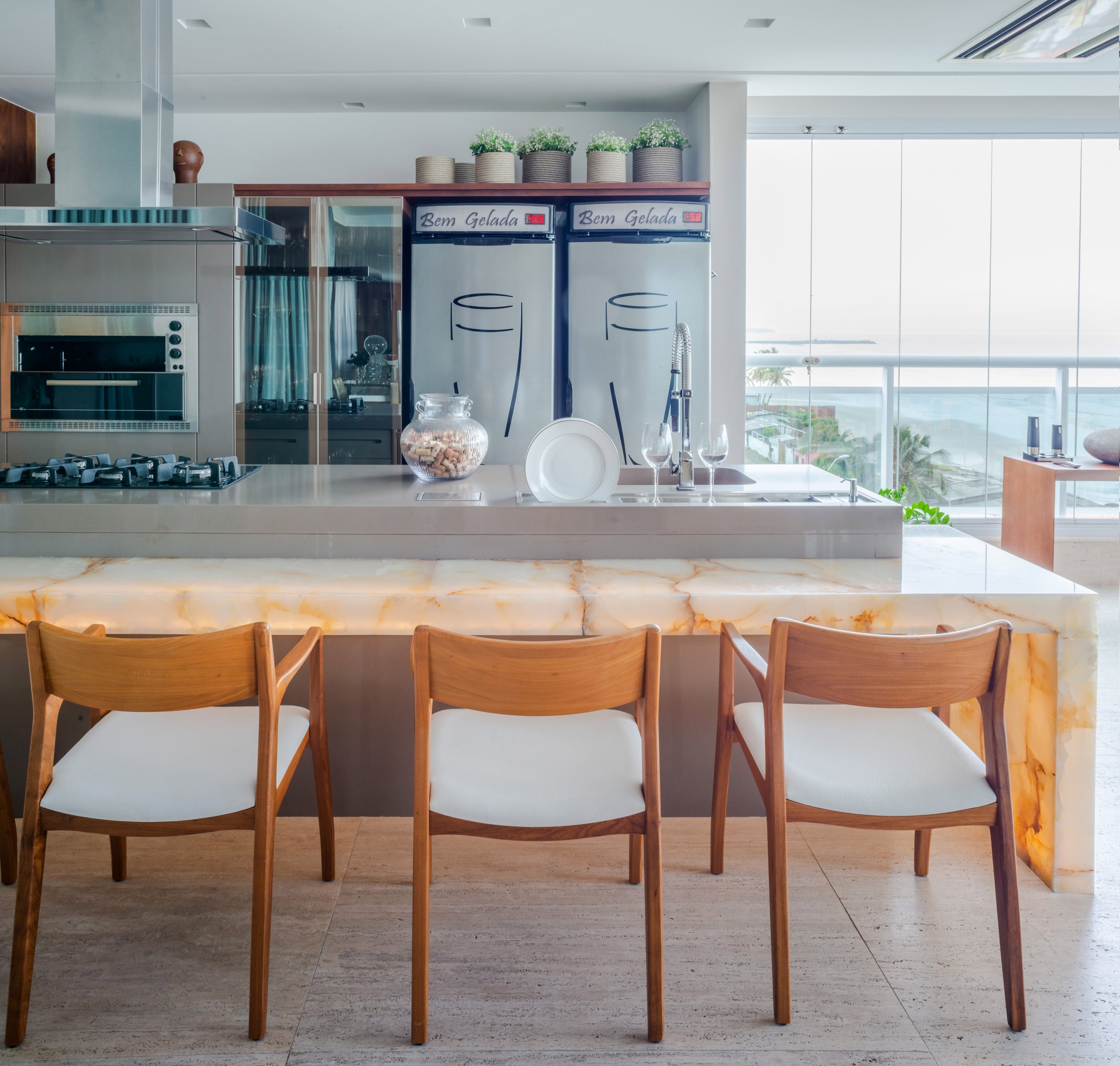 Modern kitchen with a marble island, three wooden chairs with white cushions, stainless steel appliances, potted plants on shelf, and large windows overlooking cityscape.