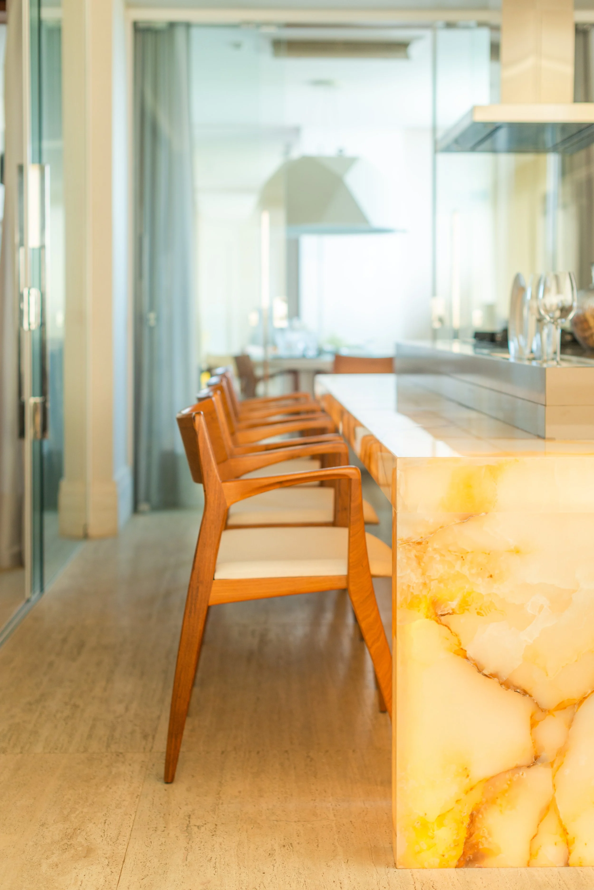 A modern kitchen with a marble island, four wooden chairs with white cushions aligned along the island, glassware on the countertop, and a large window in the background letting in natural light.