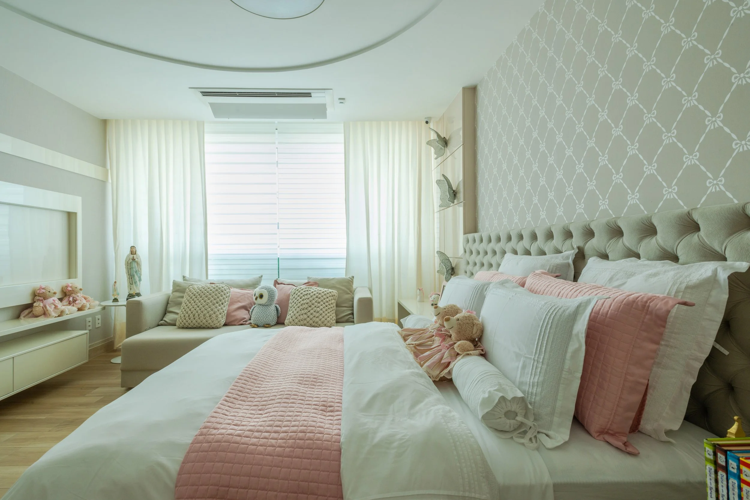 A bright and cozy bedroom with a large bed, pink quilt, and plush toys, including teddy bears and an owl, on the bed. There is a sitting area with pillows and stuffed animals near the window, which is covered with sheer white curtains. The wall behin