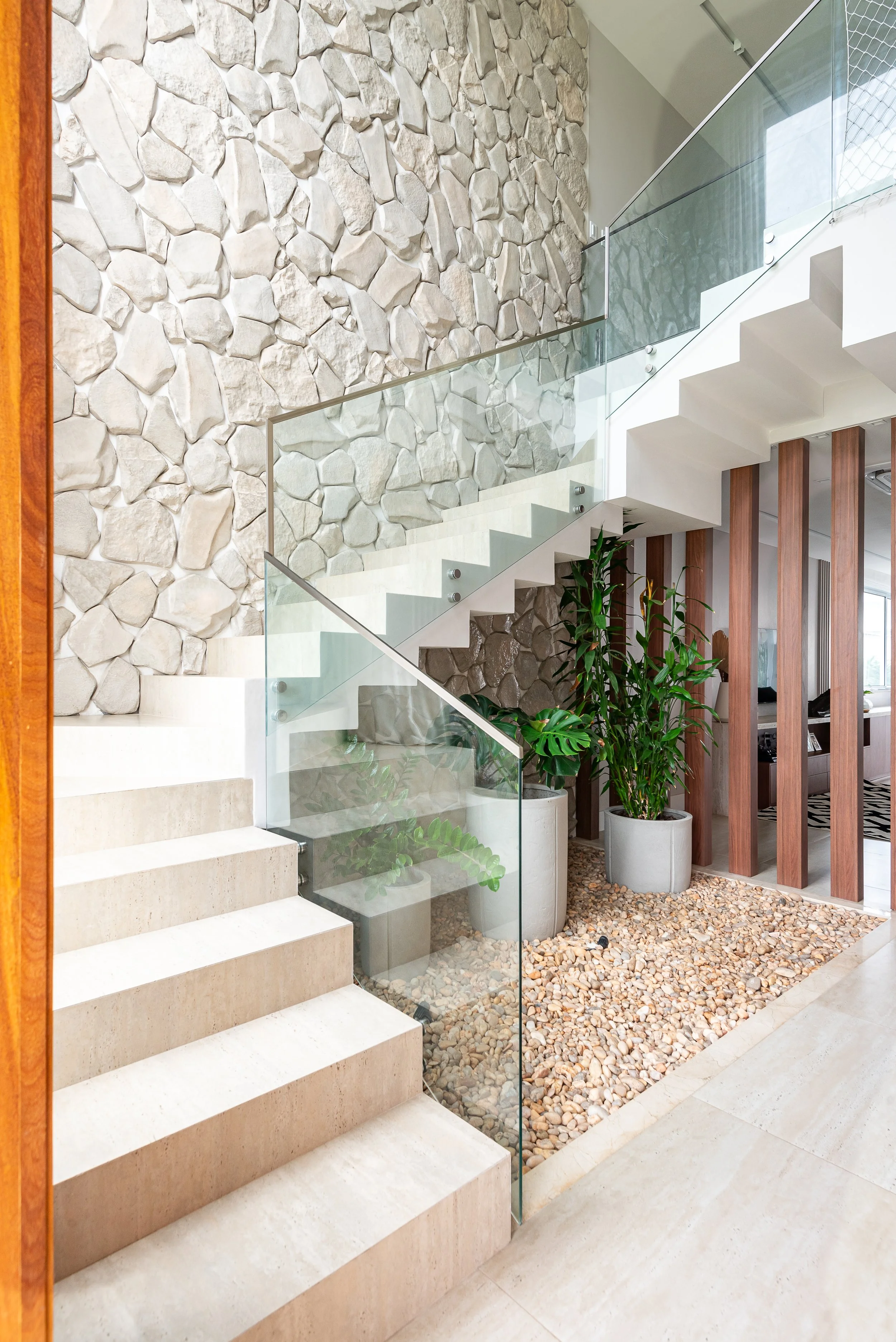 Modern interior staircase with glass railing, stone wall, potted plants, and decorative pebble ground in a contemporary home.