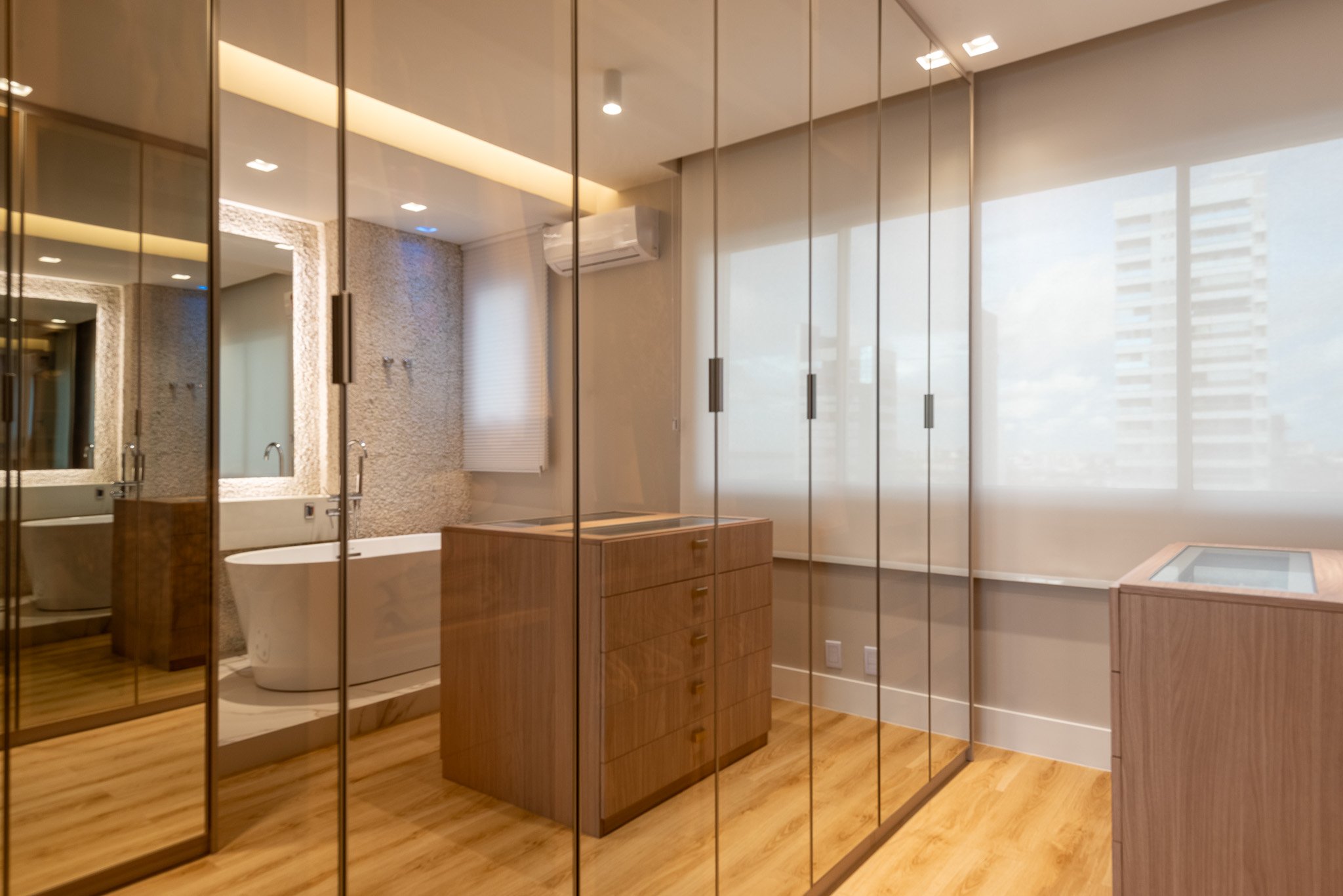 Modern bathroom with glass partition reflecting the room, featuring a bathtub, wooden furniture, and large windows with city views.