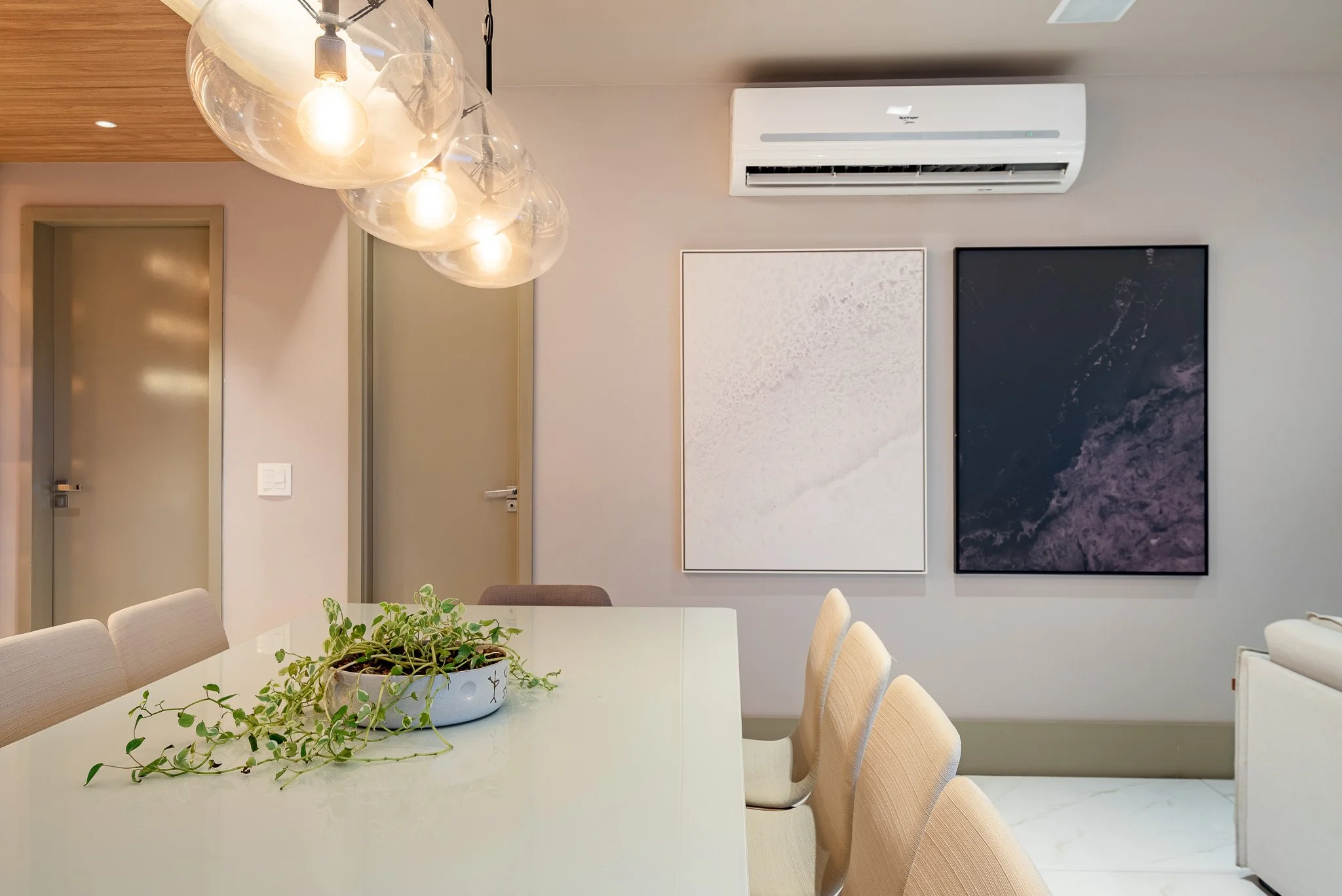 Modern dining room with a white table, beige chairs, a plant in a pot, pendant lights, wall art, and an air conditioning unit.