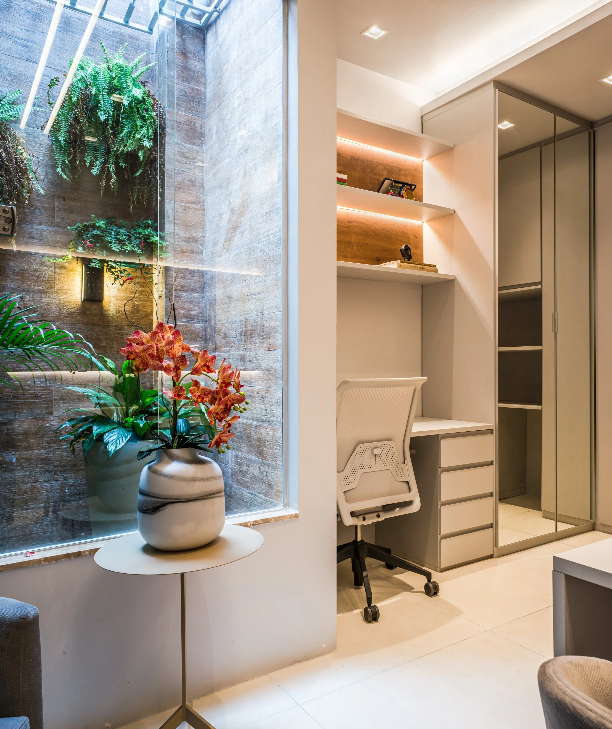 Interior of a modern room with a seating area, a white desk with a white office chair, wall-mounted shelves with books and decorative items, a closet with mirrored doors, and a large window with plants and flowers outside.