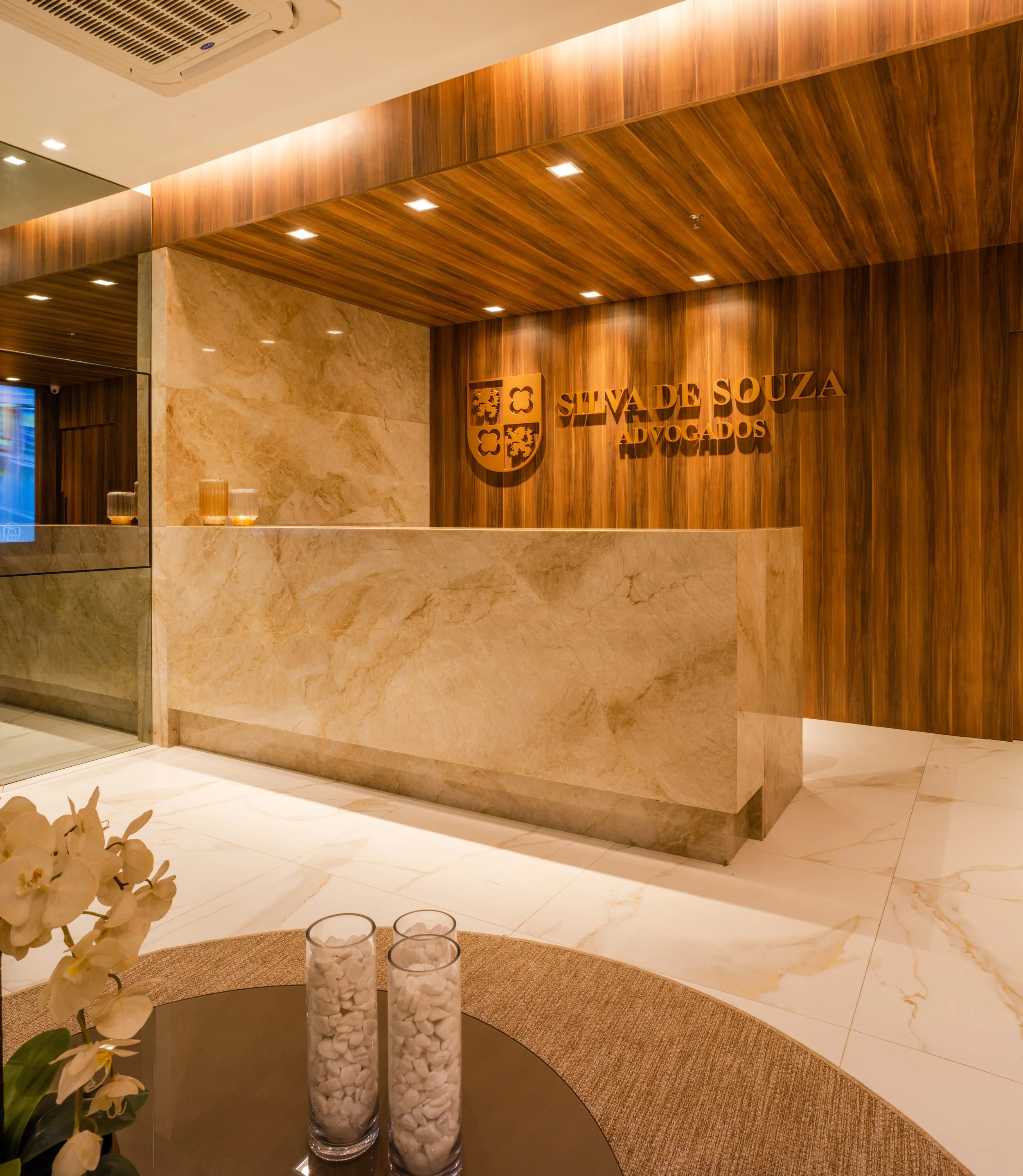 Reception area of a law firm with a wooden wall displaying the logo and name 'Silva de Souza Advogados'. A marble counter is in front, with three vases filled with white stones on a round table nearby. Flowers are partially visible in the foreground.