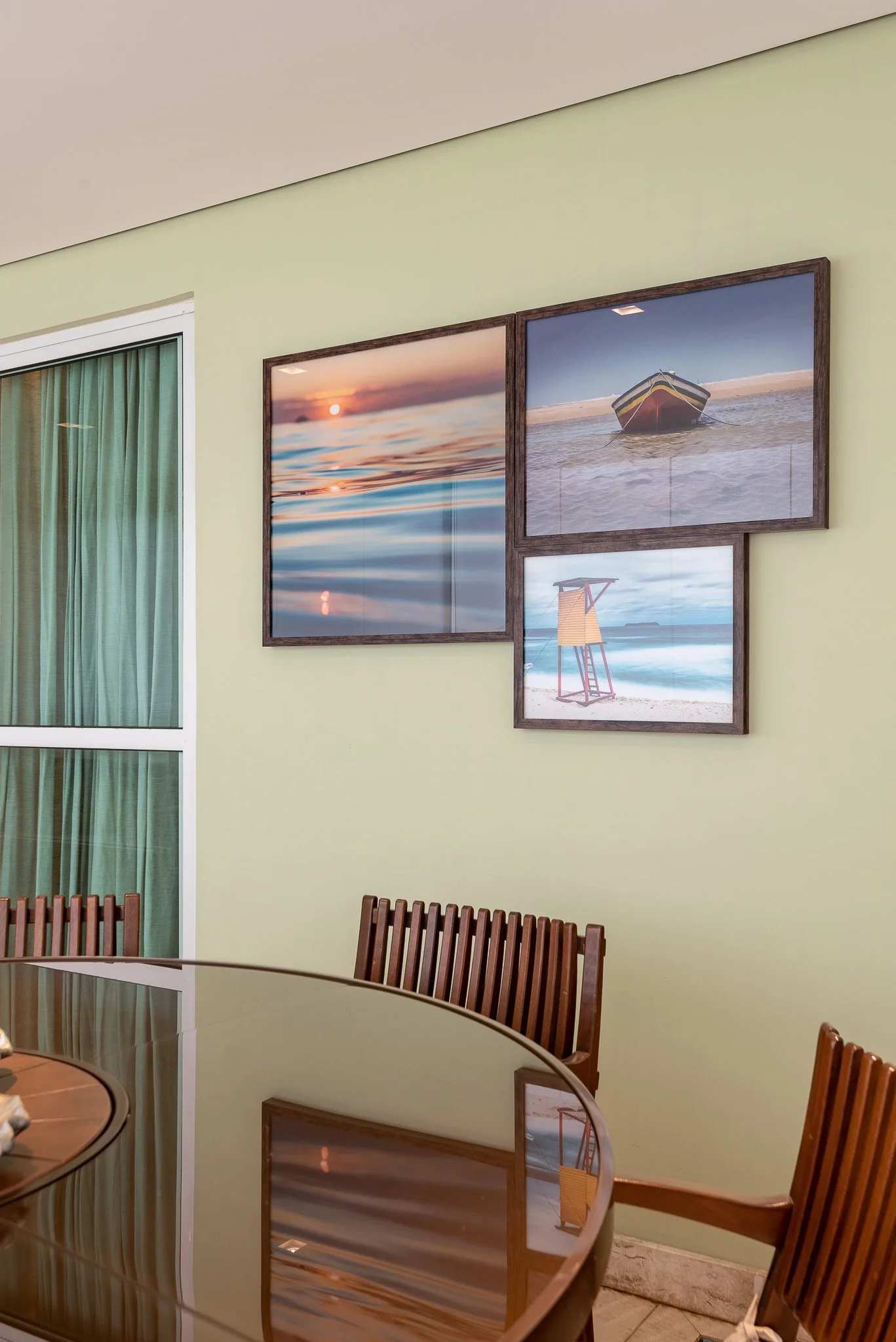 Interior of a dining area with a polished wooden table and wooden chairs. Decorated wall features three framed pictures of a sunset over water, a boat on the beach, and a lifeguard tower on the sand. A window with green curtains is visible on the lef