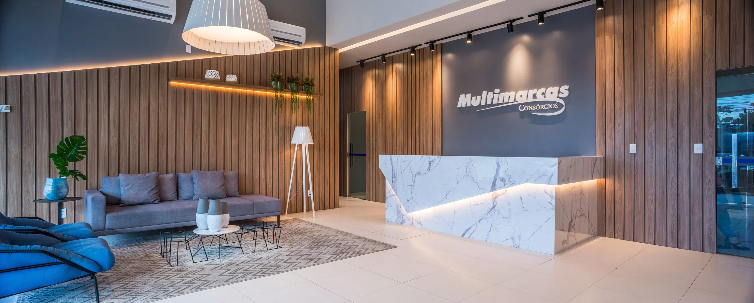 Modern office reception area with a marble front desk, wooden wall paneling, a gray sofa with cushions, a floor lamp, decorative vases, and a wall sign that reads 'Multimarcas Consórcios'.