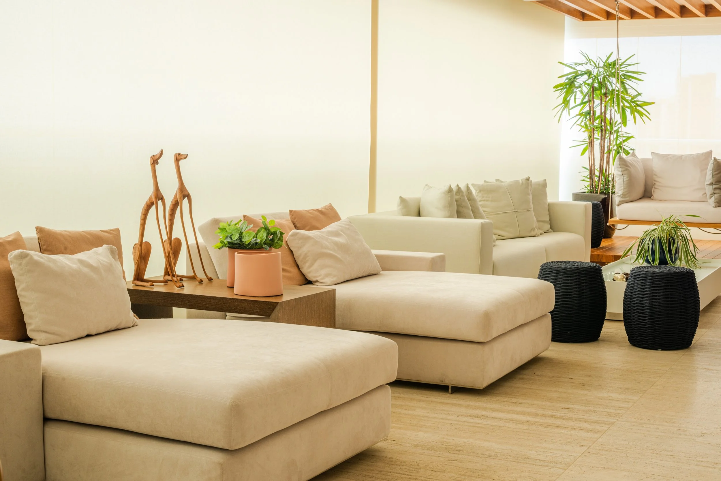 Living room with beige sofas, black woven stools, potted plants, and wooden giraffe sculptures.