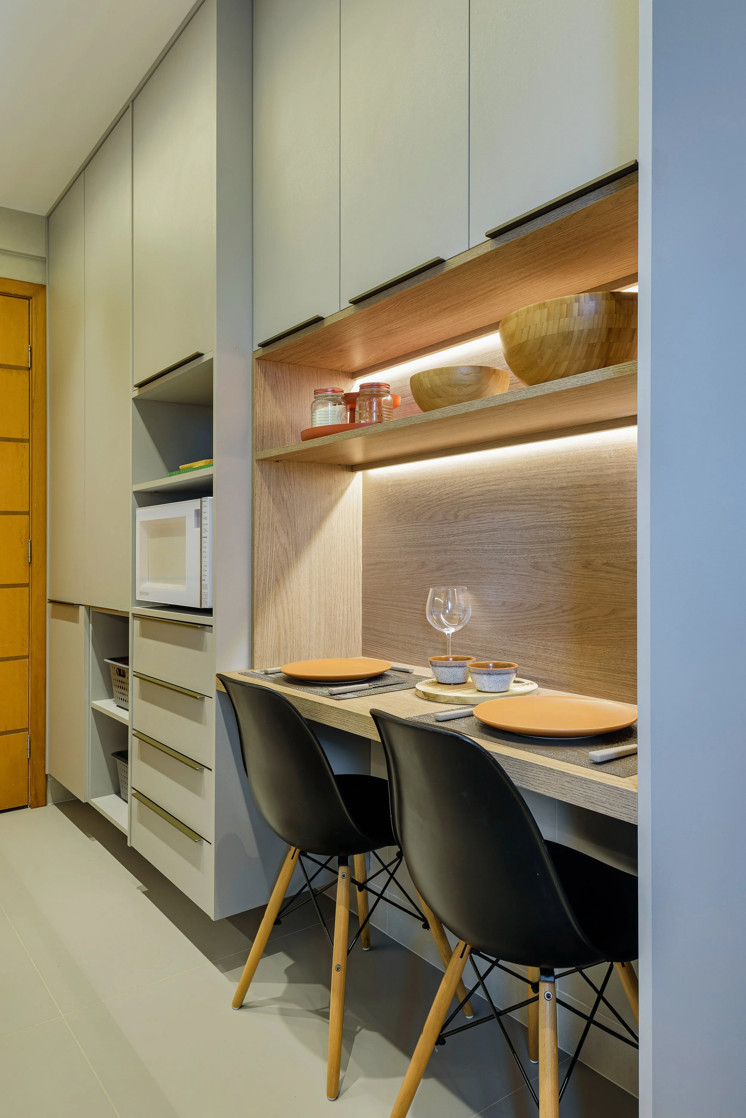 Modern kitchen nook with two black chairs, a built-in wooden counter with place settings, two shelves with wooden bowls and jars, and a surrounding wall of white cabinets.