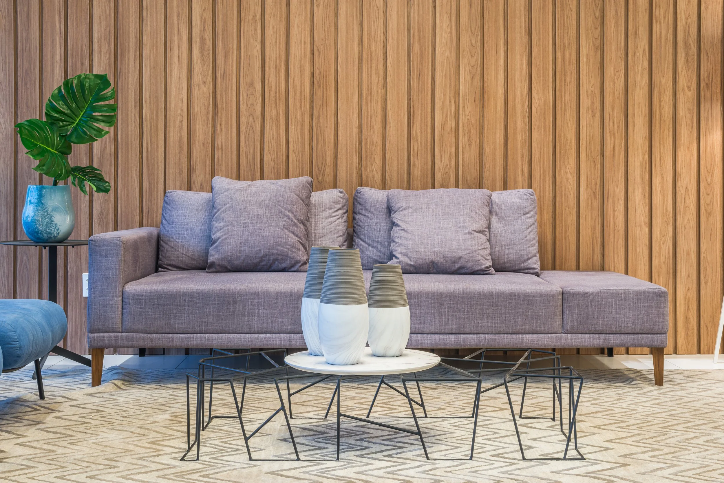 Living room with a grey sofa, light grey pillows, a wooden paneled wall, a side table with a green leafy plant in a blue vase, a round coffee table with three decorative vases, and a light patterned carpet.