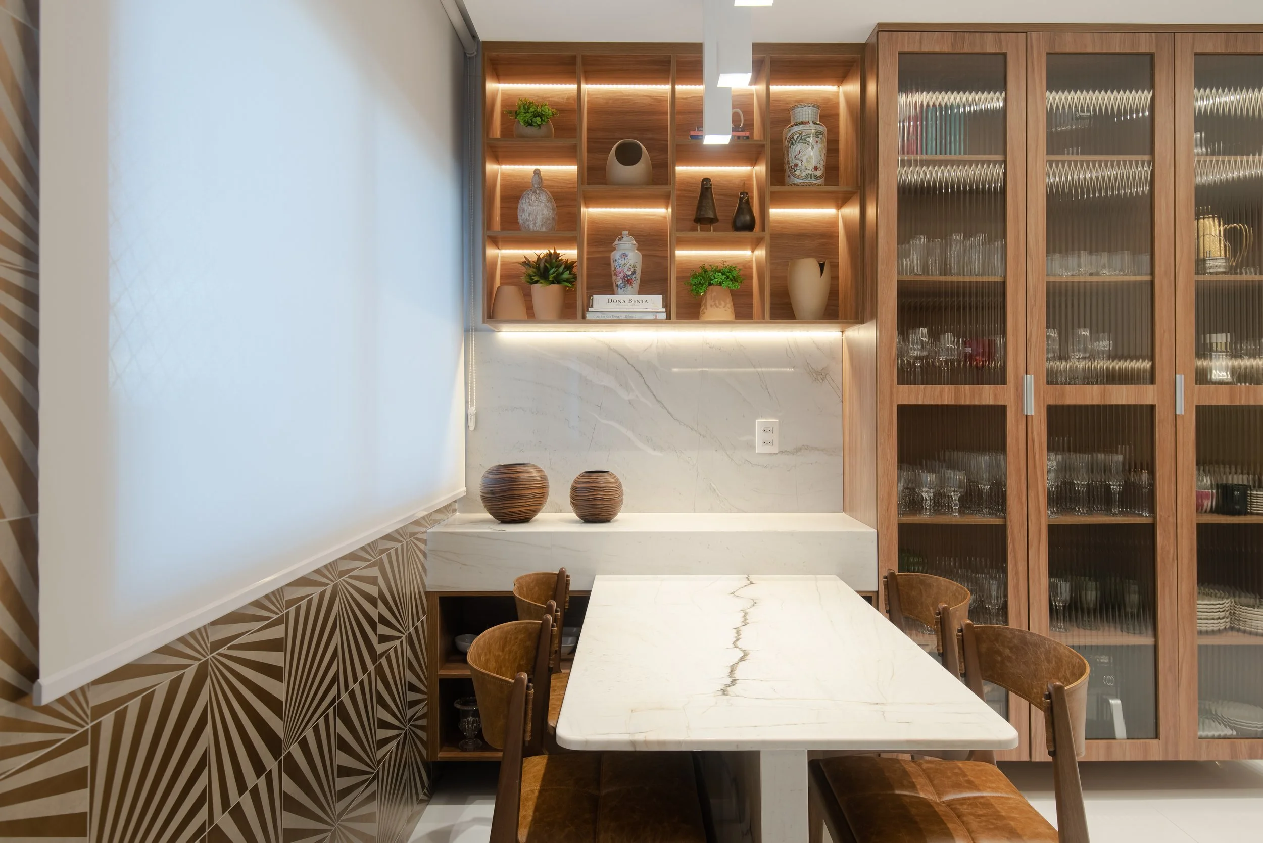 Modern dining area with a white marble table, wooden chairs, and a built-in cabinet with glass doors containing glassware. A wall-mounted shelving unit with decorative vases, plants, and books is lit with warm lighting, and a window with a white roll