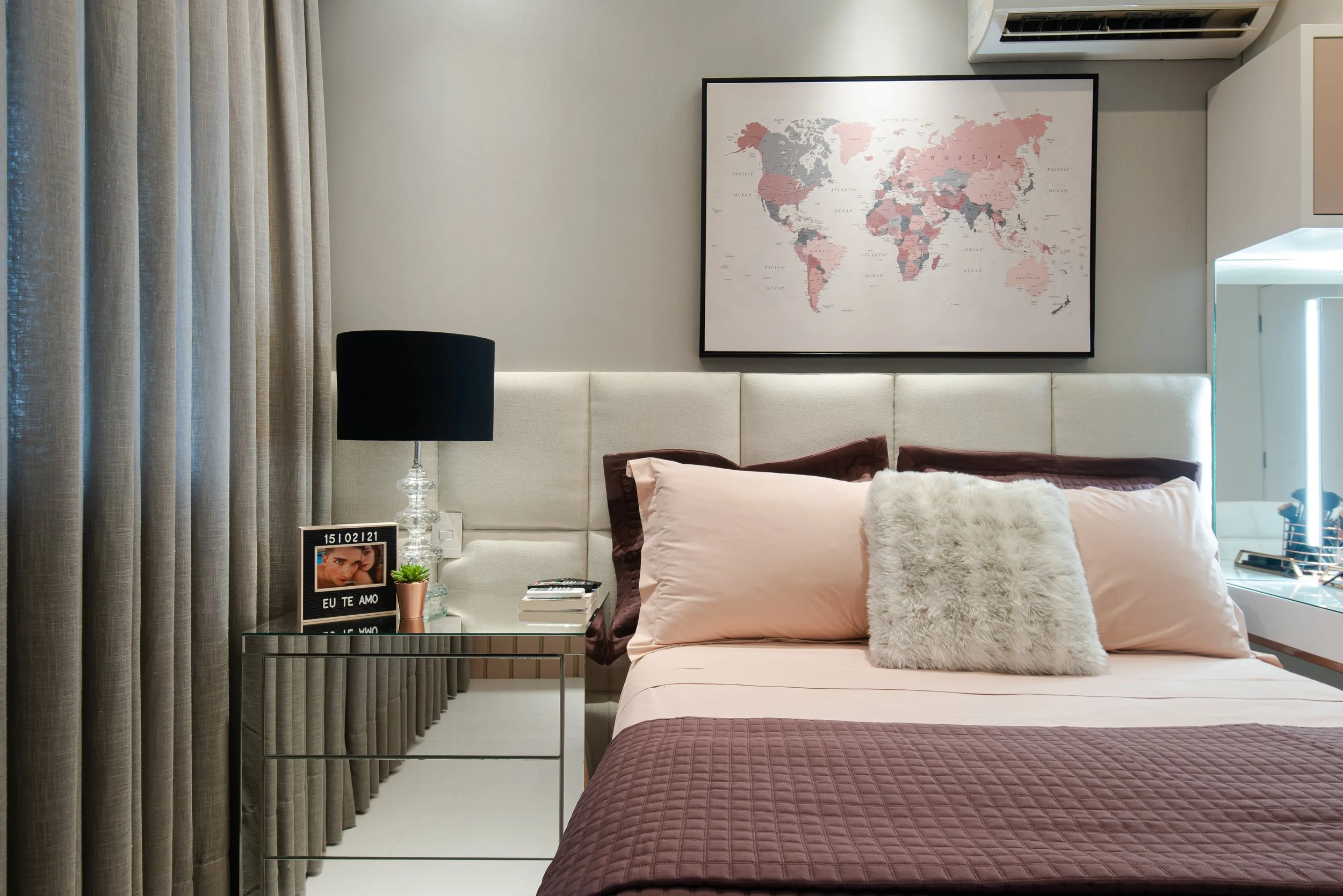 A bedroom with a bed, pillows, a black table lamp, a framed photo with the text 'EU TE AMO,' a small potted plant, a world map on the wall, beige curtains, and a mirrored dresser.