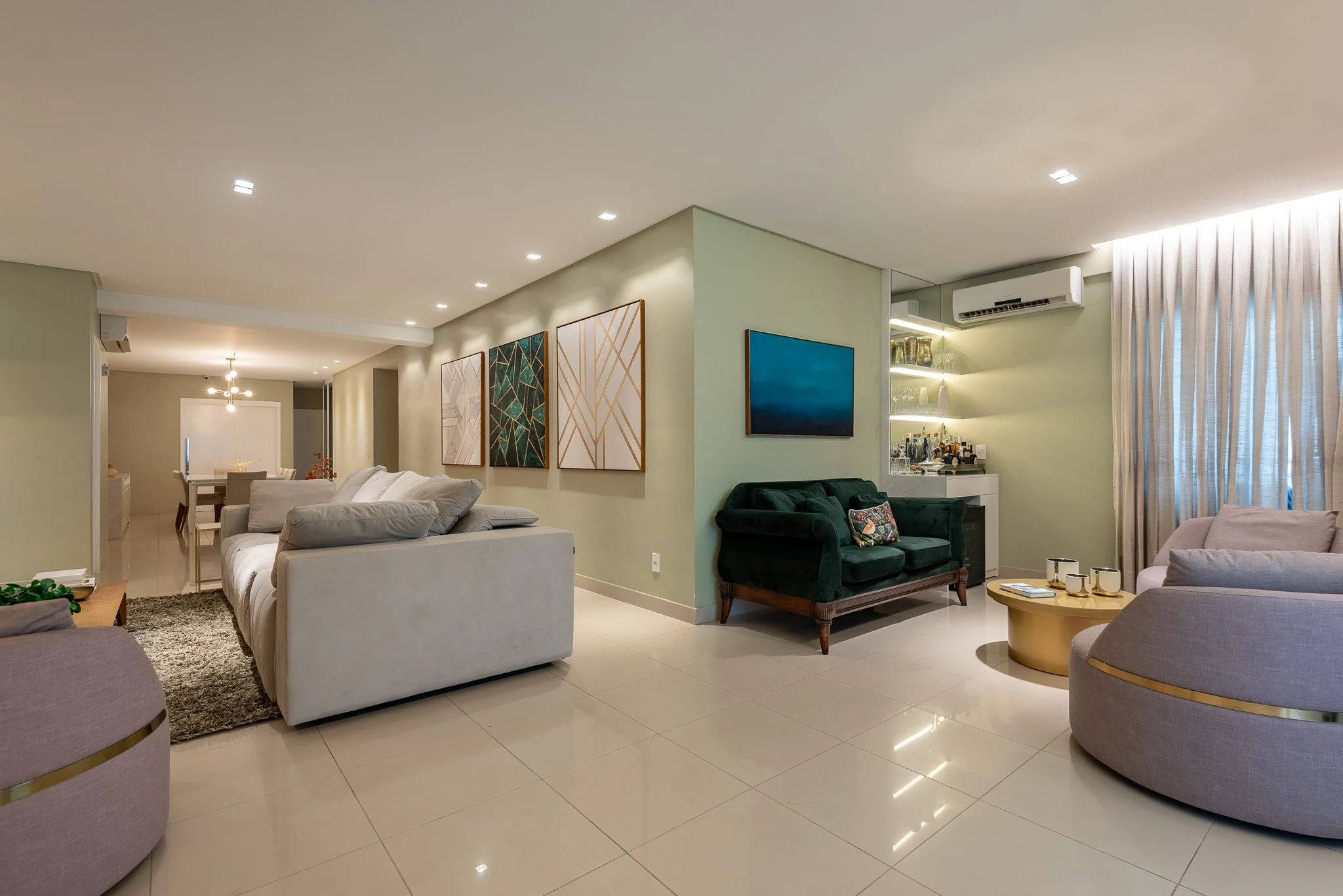 Modern living room with white sofa, green velvet loveseat, circular purple chair, dining area in background, art on walls, bar area with shelves and bottles, beige tiled floor, large window with curtains, and ceiling lights.