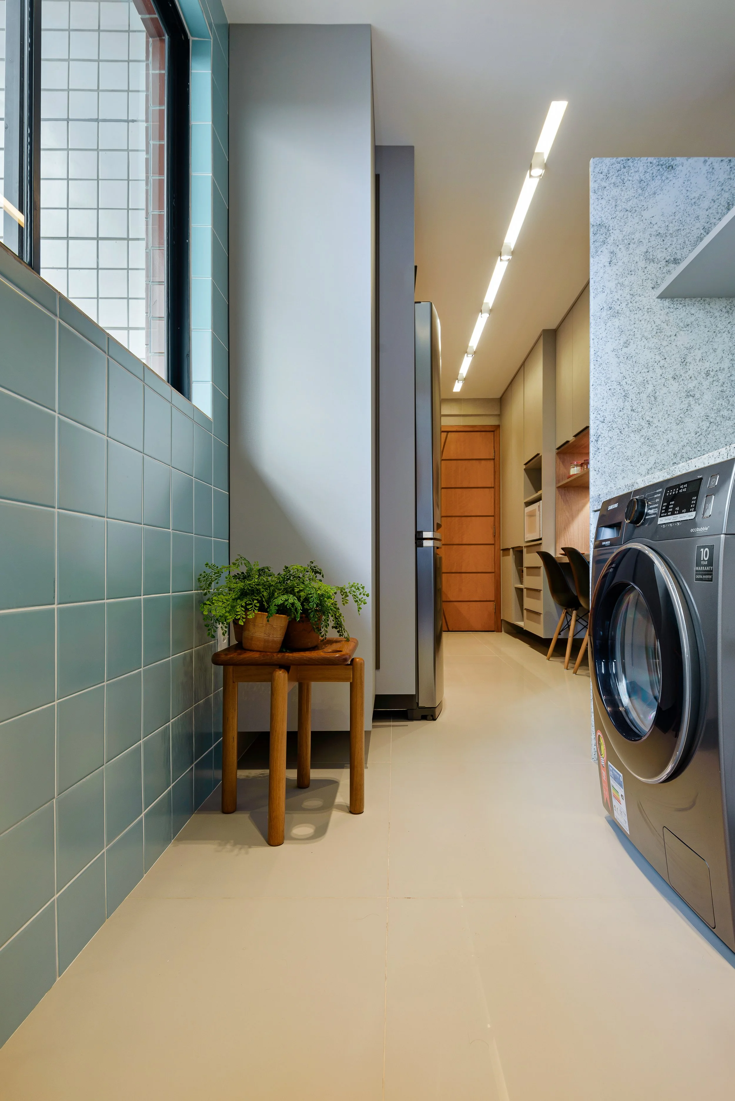 Laundry room with light beige floor, teal tiled wall, large window, wooden stool with potted plant, stainless steel washing machine, built-in cabinets, and a wooden door at the back.