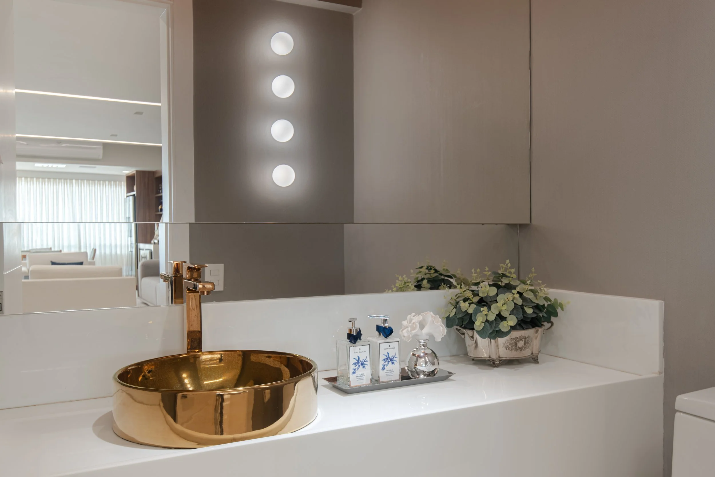 Bathroom sink with a gold basin, gold faucet, and a silver tray holding soap dispensers and a small vase with white flowers. There is a plant in a decorative pot on the counter, and a mirror reflecting a living room with white furniture and large win