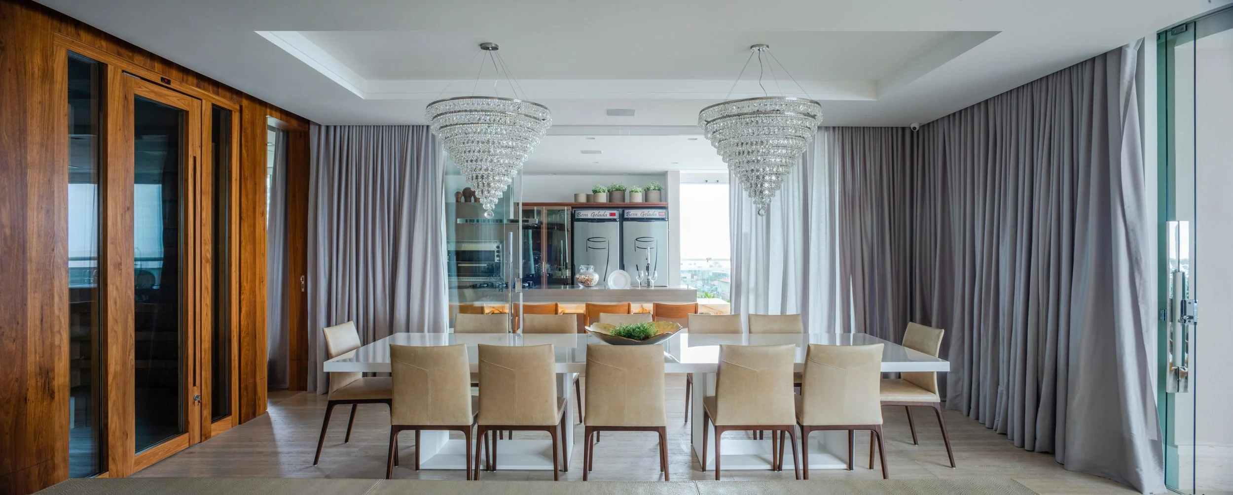 Modern dining room with a large white table, cream chairs, wooden paneling, and two large crystal chandeliers, with floor-to-ceiling curtains and sliding glass door.