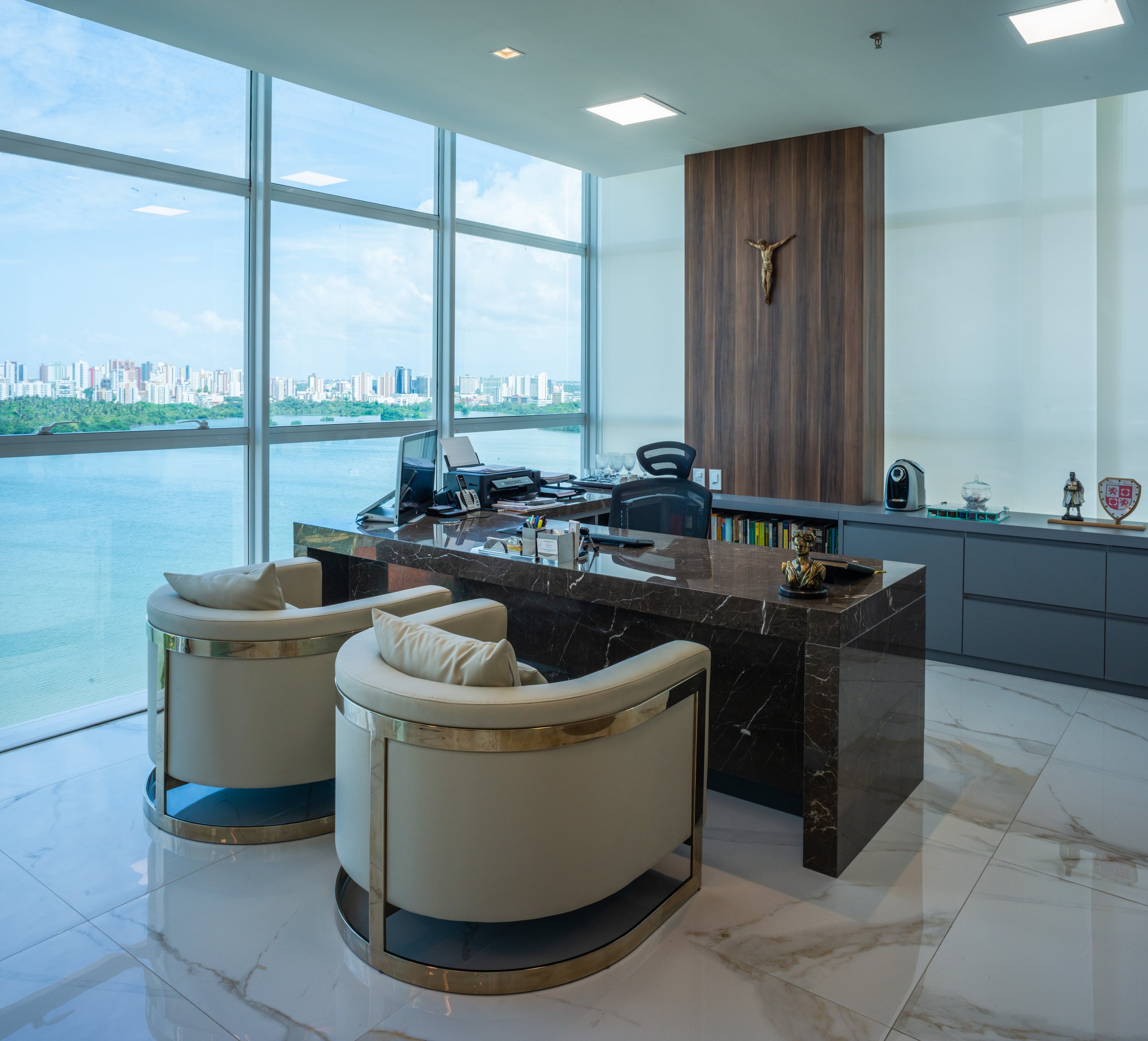 Modern office with large windows offering a city skyline view, featuring a dark marble desk, two cream-colored chairs, and decorative items including a crucifix on a wooden wall panel.