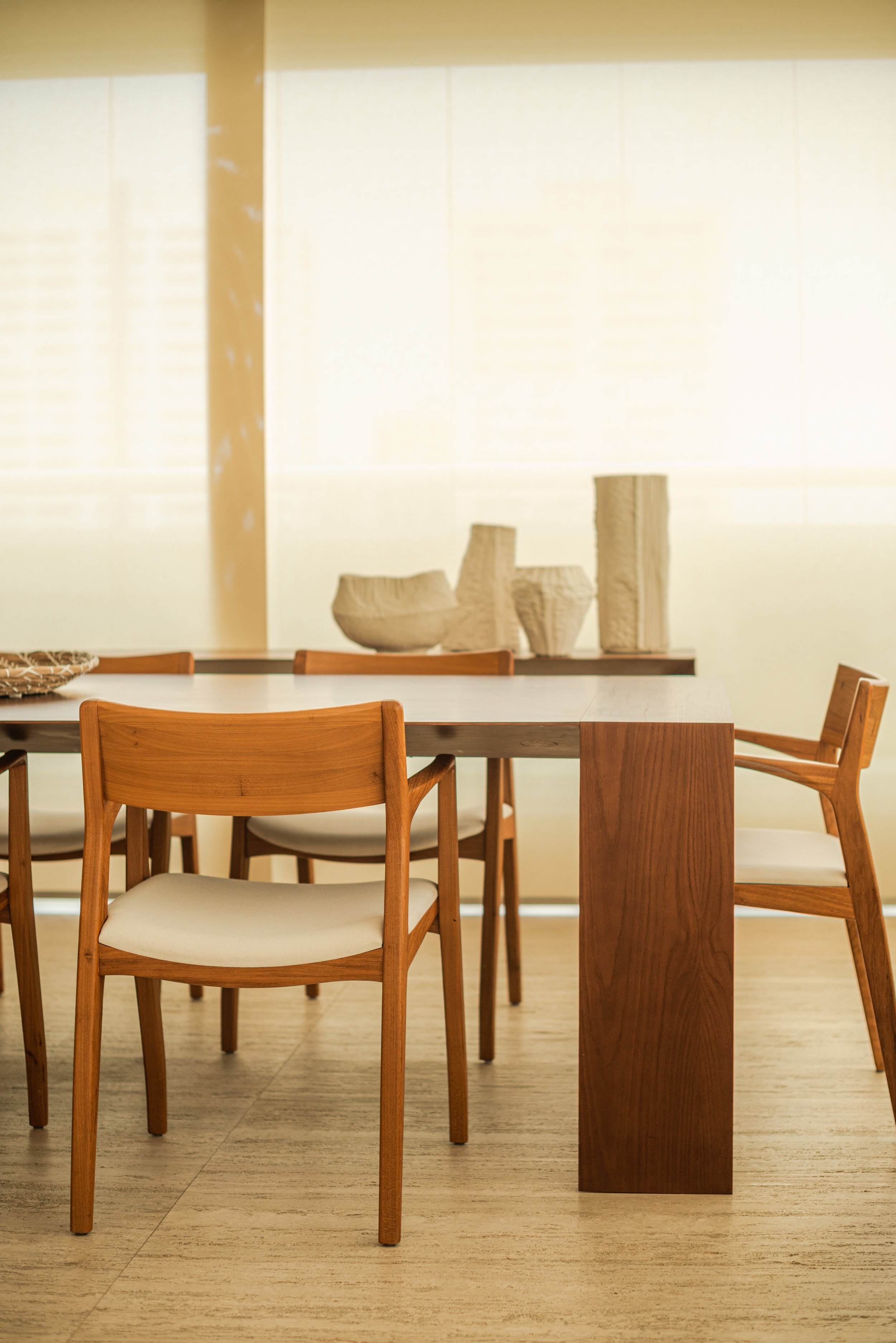 Wooden dining table with six wooden chairs in a well-lit room with beige curtains and ceramic vases on a shelf in the background.