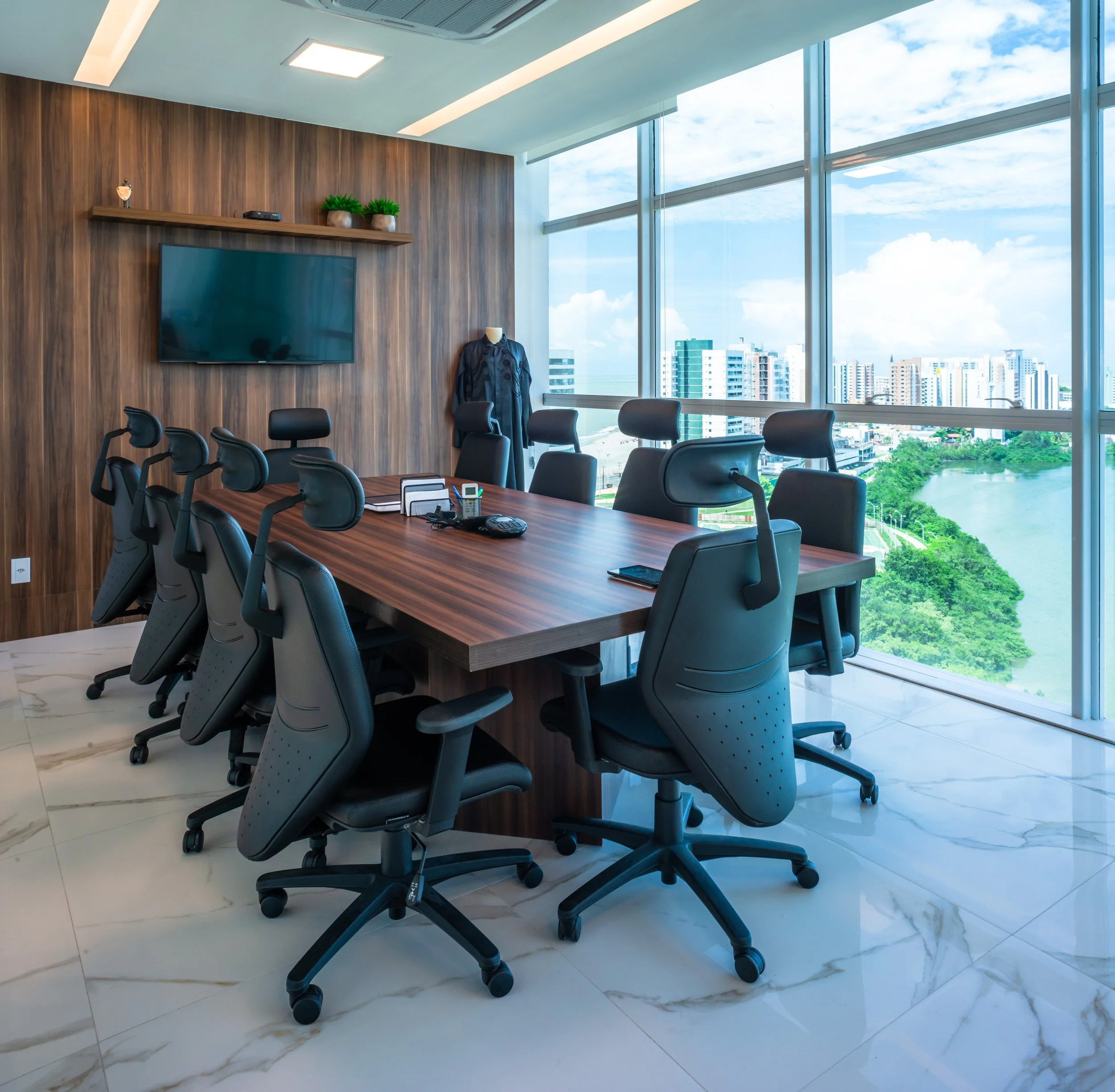 Modern conference room with a large wooden table and black office chairs, floor-to-ceiling windows showing a cityscape and river, a wall-mounted TV, and minimal decor.