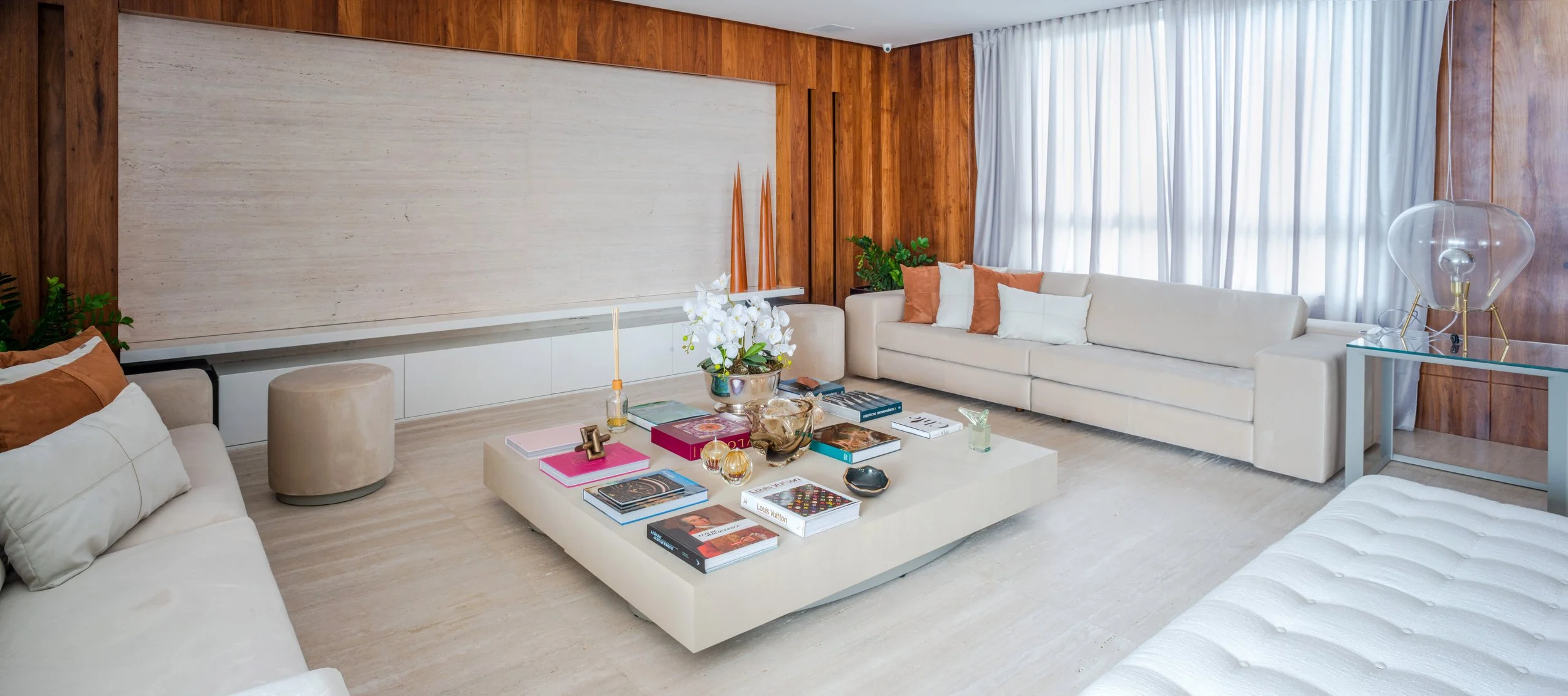 Modern living room with white sofa, coffee table with books and decorations, large window with curtains, wooden wall accents, and decorative lamps.