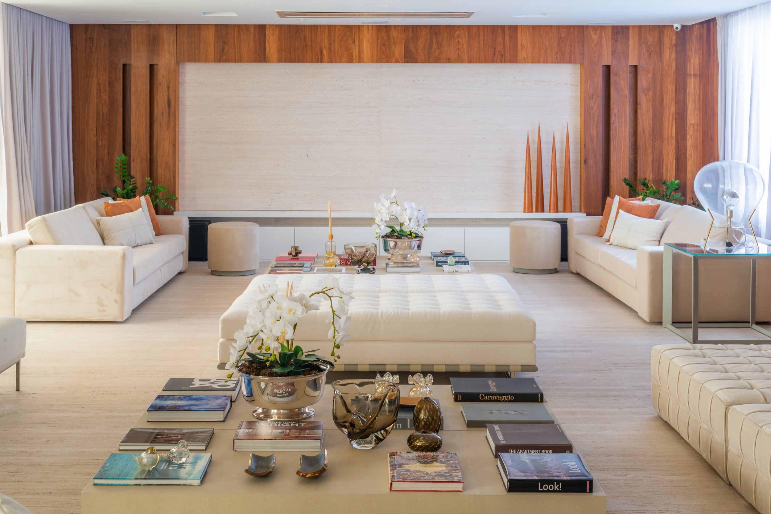 A spacious modern living room with white sofas, a large central ottoman, decorative books, vases, and plants, and a wood-paneled wall with contemporary art and sculptures.