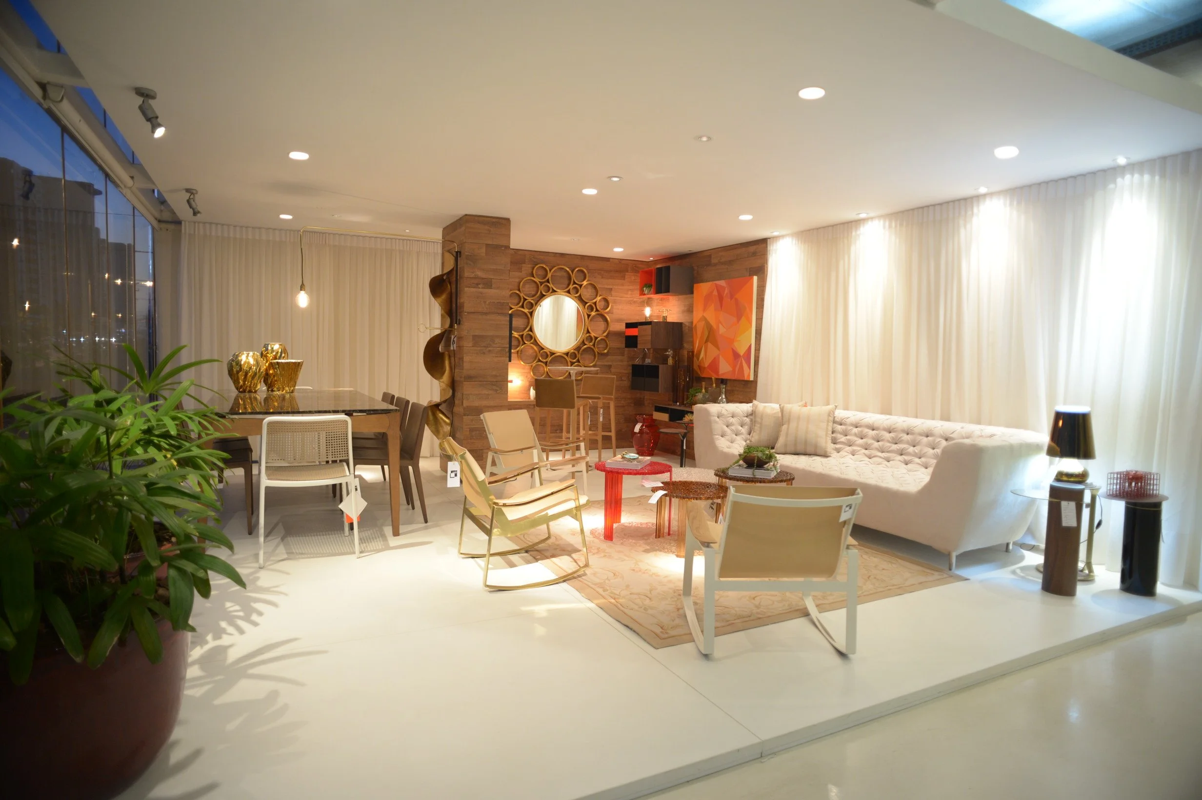 Modern living room with various furniture pieces including a white tufted sofa, wooden chairs, and a dining table. Decor includes gold vases, a large round mirror with a bow-shaped frame, colorful abstract artwork, and a potted plant. The space is il