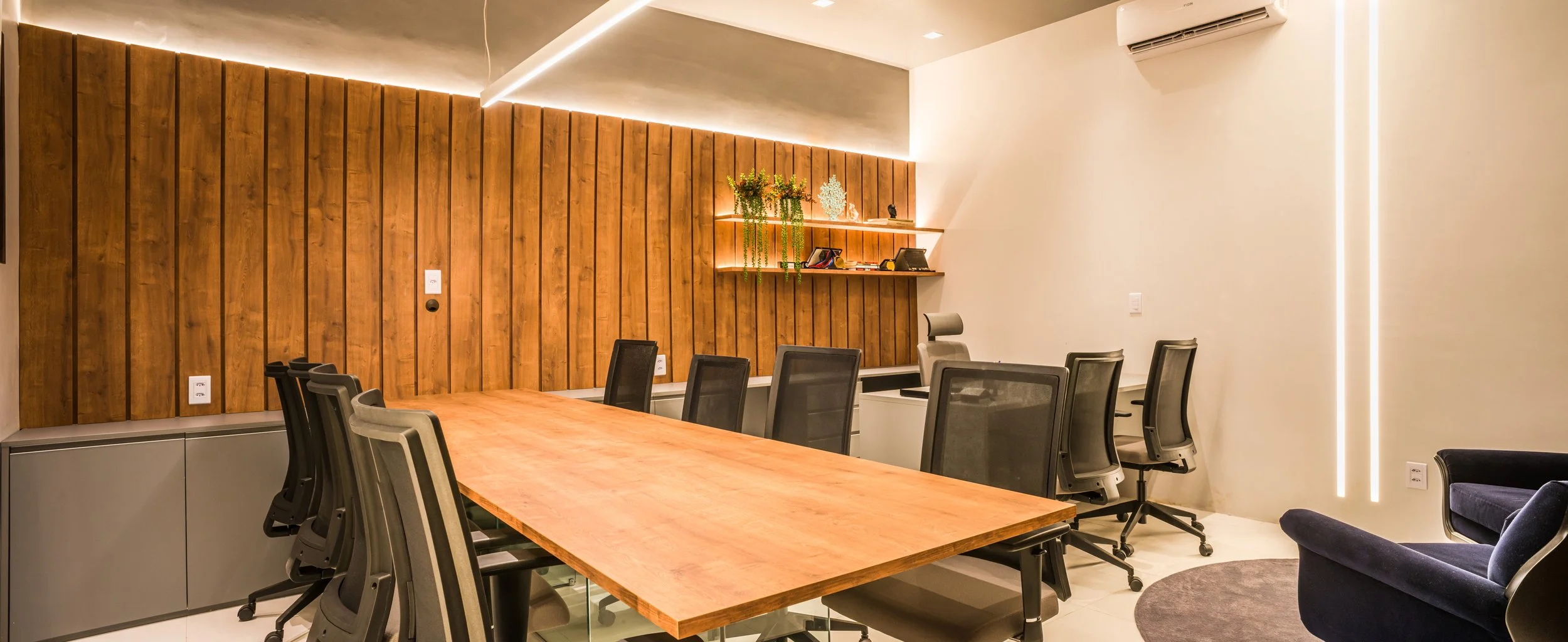 Modern conference room with a large wooden table surrounded by black office chairs, a wooden accent wall with shelves, and a white wall with vertical lighting.