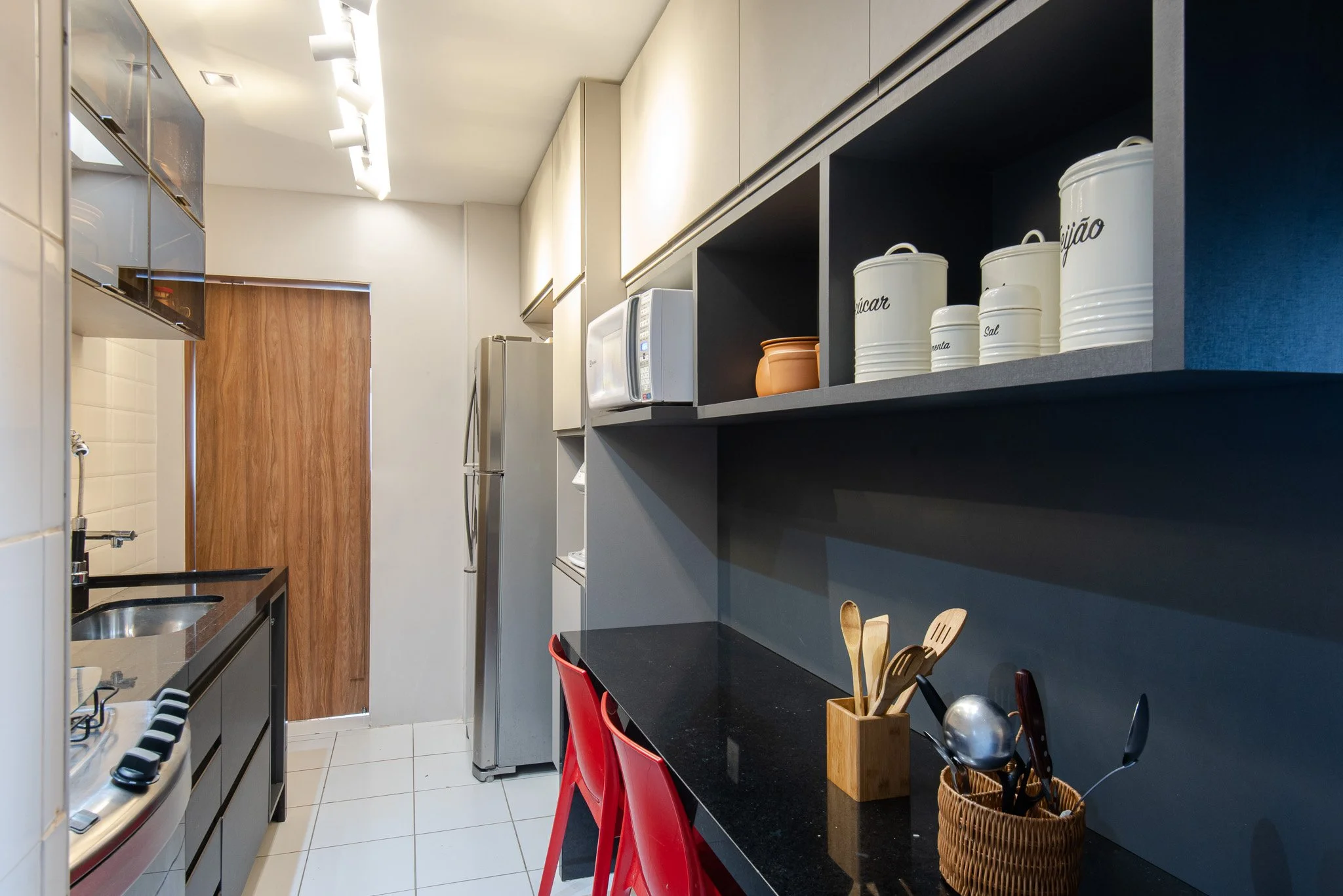 Small modern kitchen with white tiled floor and dark countertops, red chairs, wooden wall, and open shelves with labeled jars and containers.