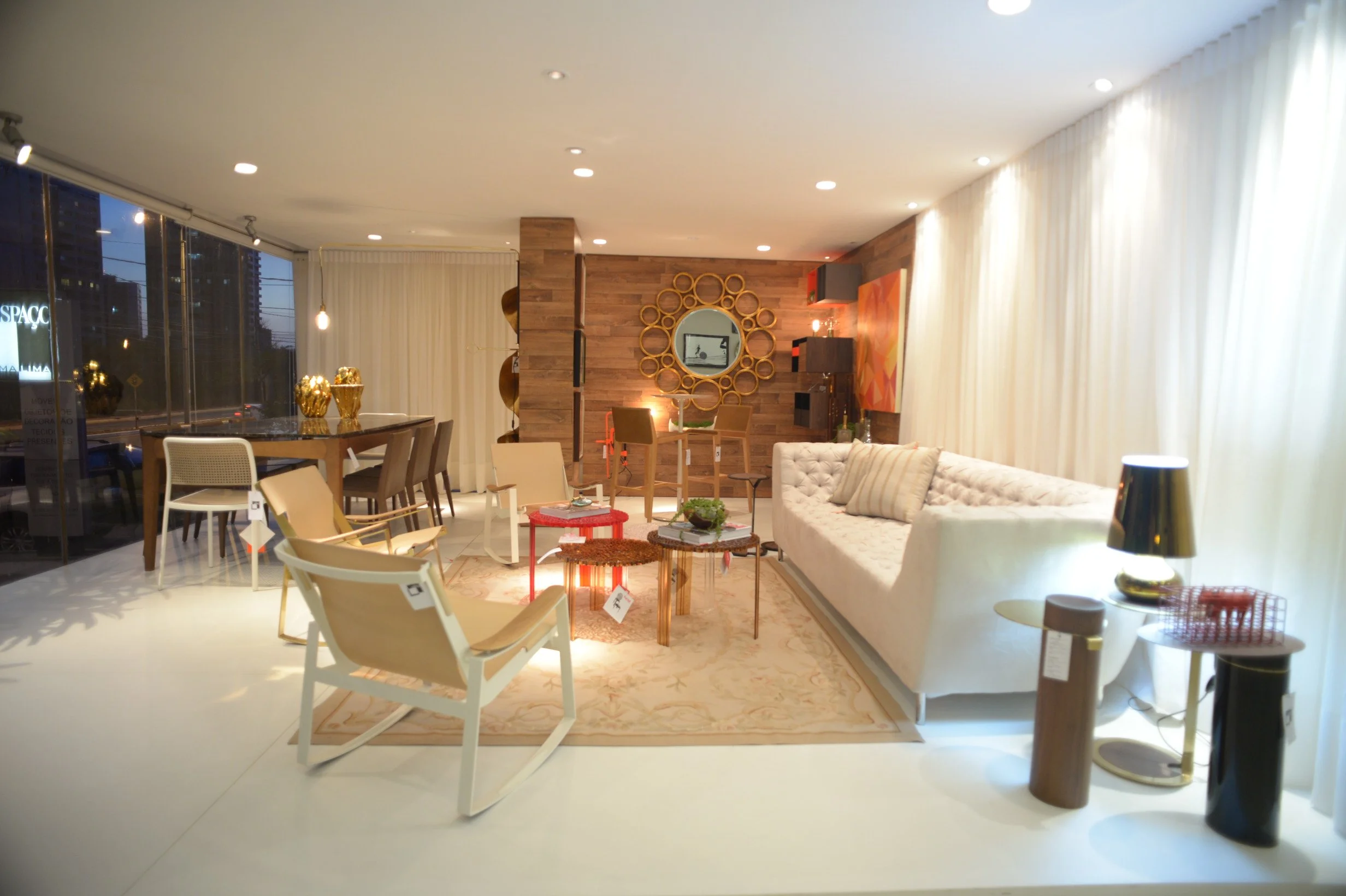 Modern living room with white tufted sofa, wooden wall with circular mirror, and various chairs around coffee tables, illuminated by ceiling and table lamps, with large windows and white curtains.