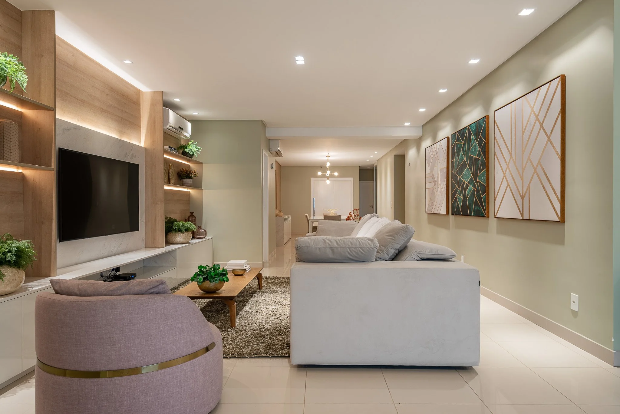 Modern living room with cream sofa, purple armchair, wooden coffee table, wall-mounted TV, built-in shelves with plants, artwork on the wall, and an open dining area in the background.