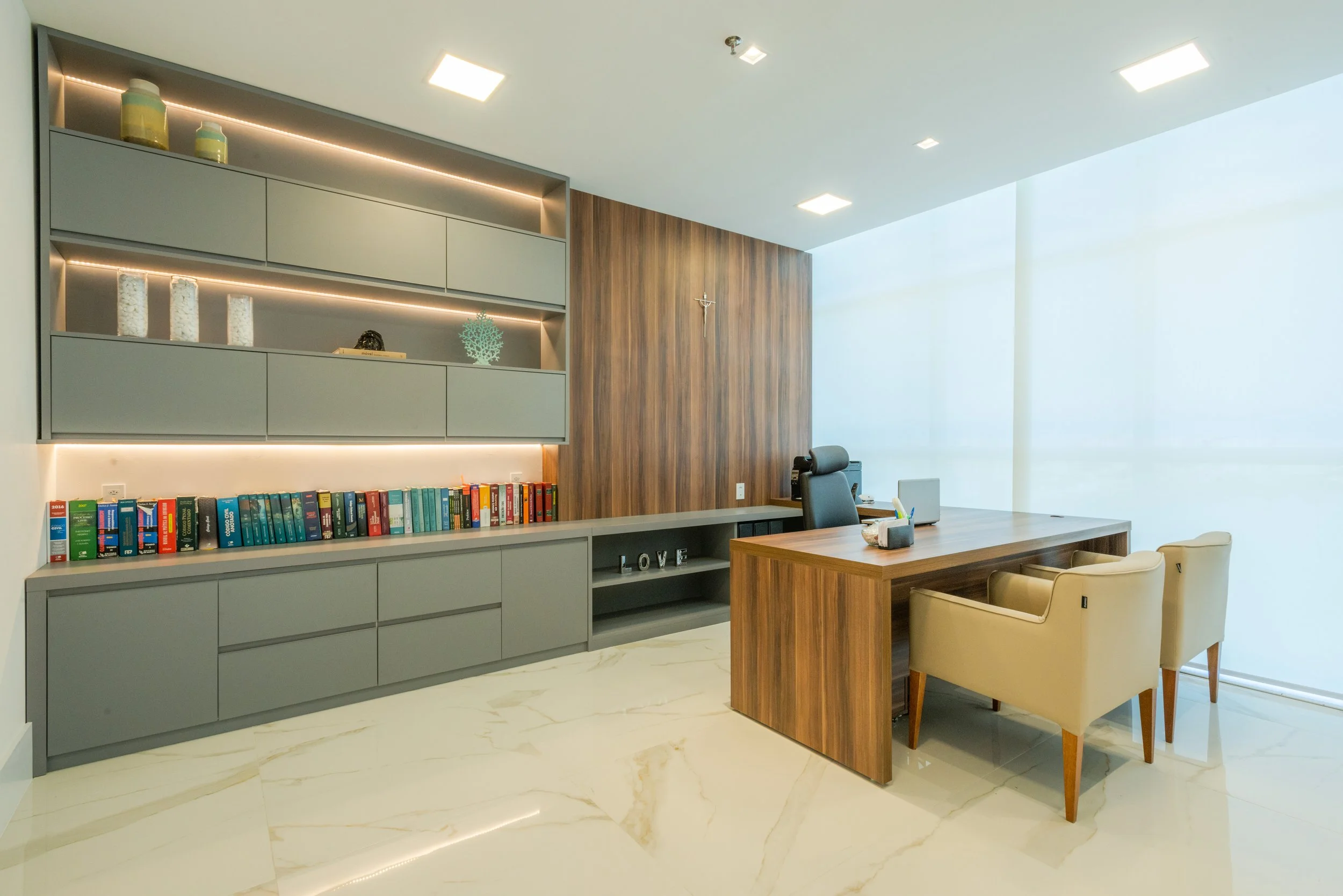 Modern office with a large wooden desk, beige chairs, gray wall shelving with books and decorative items, and a wooden accent wall with a crucifix, illuminated by ceiling lights and large window.