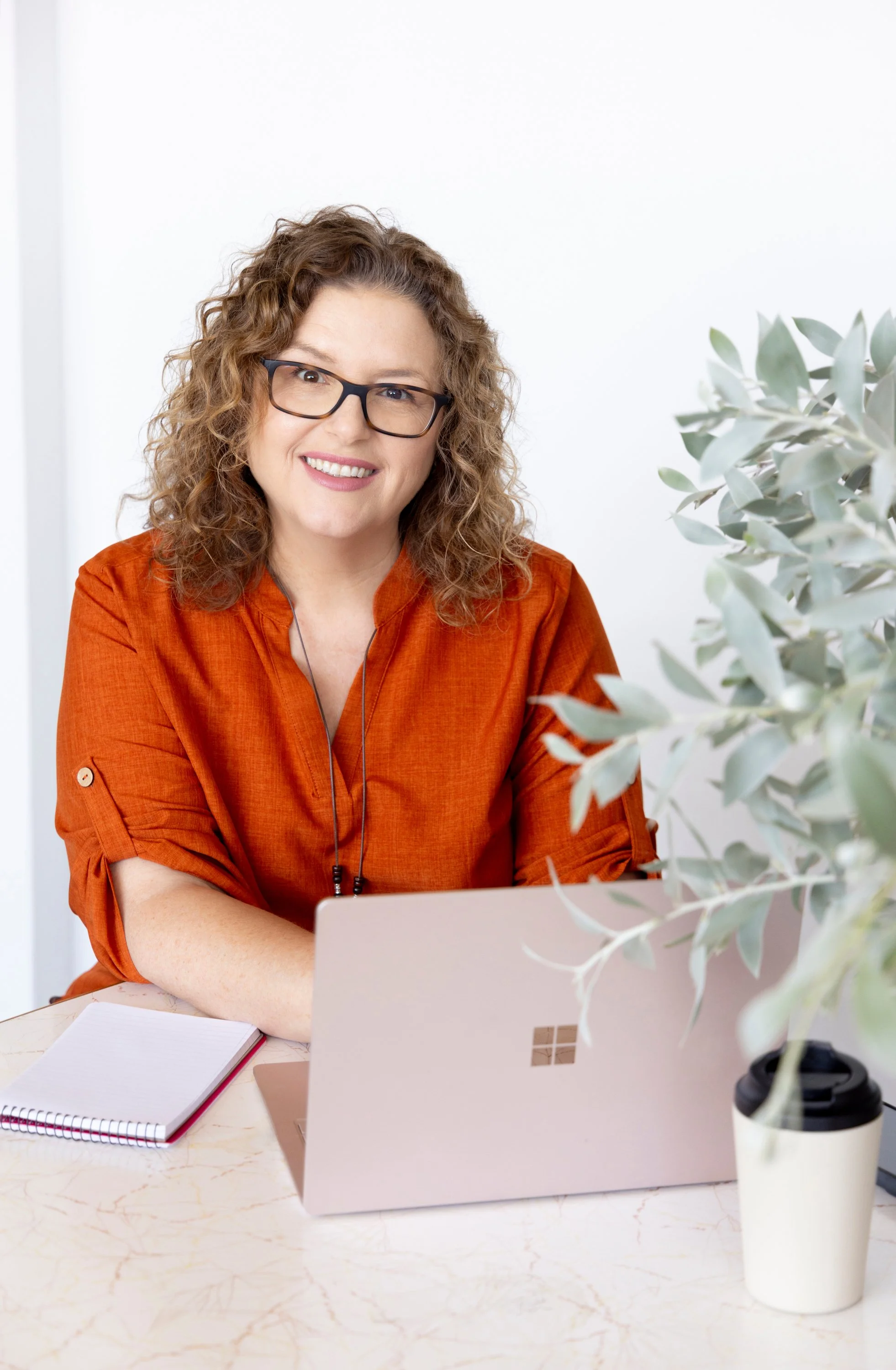 A woman with curly hair and glasses sitting at a desk, smiling, in front of a silver Microsoft Surface laptop, with a notebook to her left, and a cup of coffee to her right .Lee Sailsbury, Pool & Spa industry coach