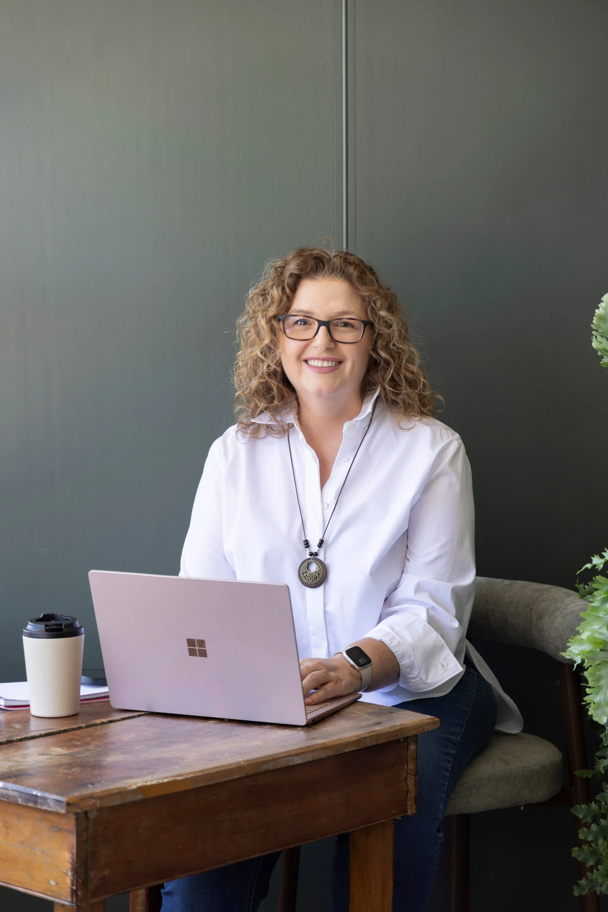 Pool & Spa business coach. A woman with curly hair and glasses sitting at a wooden table with a laptop, coffee cup, and a plant nearby, smiling at the camera.