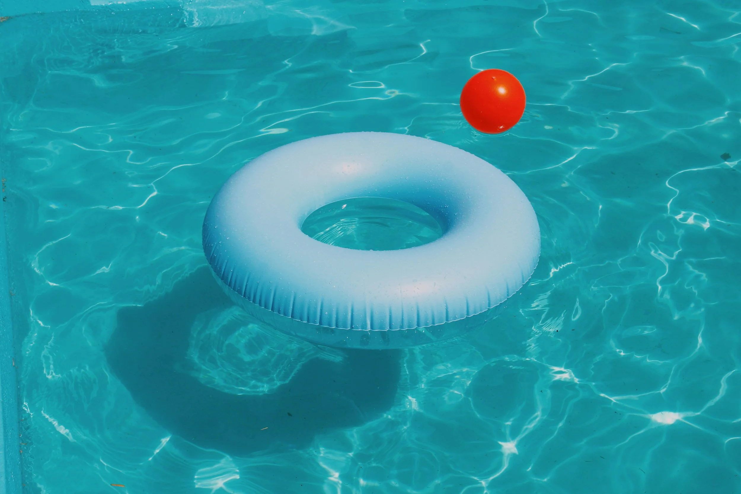 A white inflatable donut float and a red ball floating in a swimming pool with clear blue water.