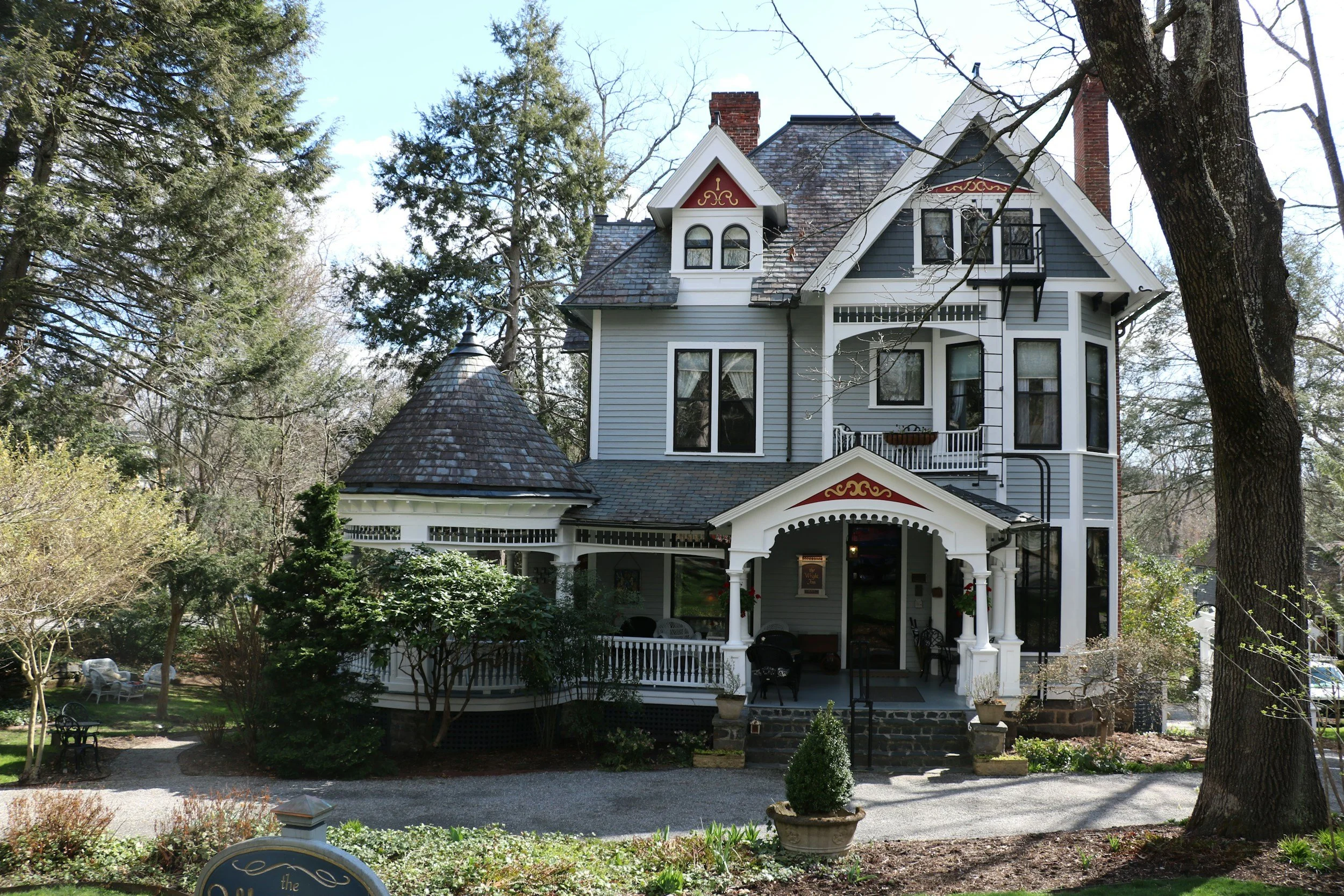 A Victorian-style house with a gray exterior, multiple gables, bay windows, and a porch. The house is surrounded by trees and has a garden with benches and plants in the yard.