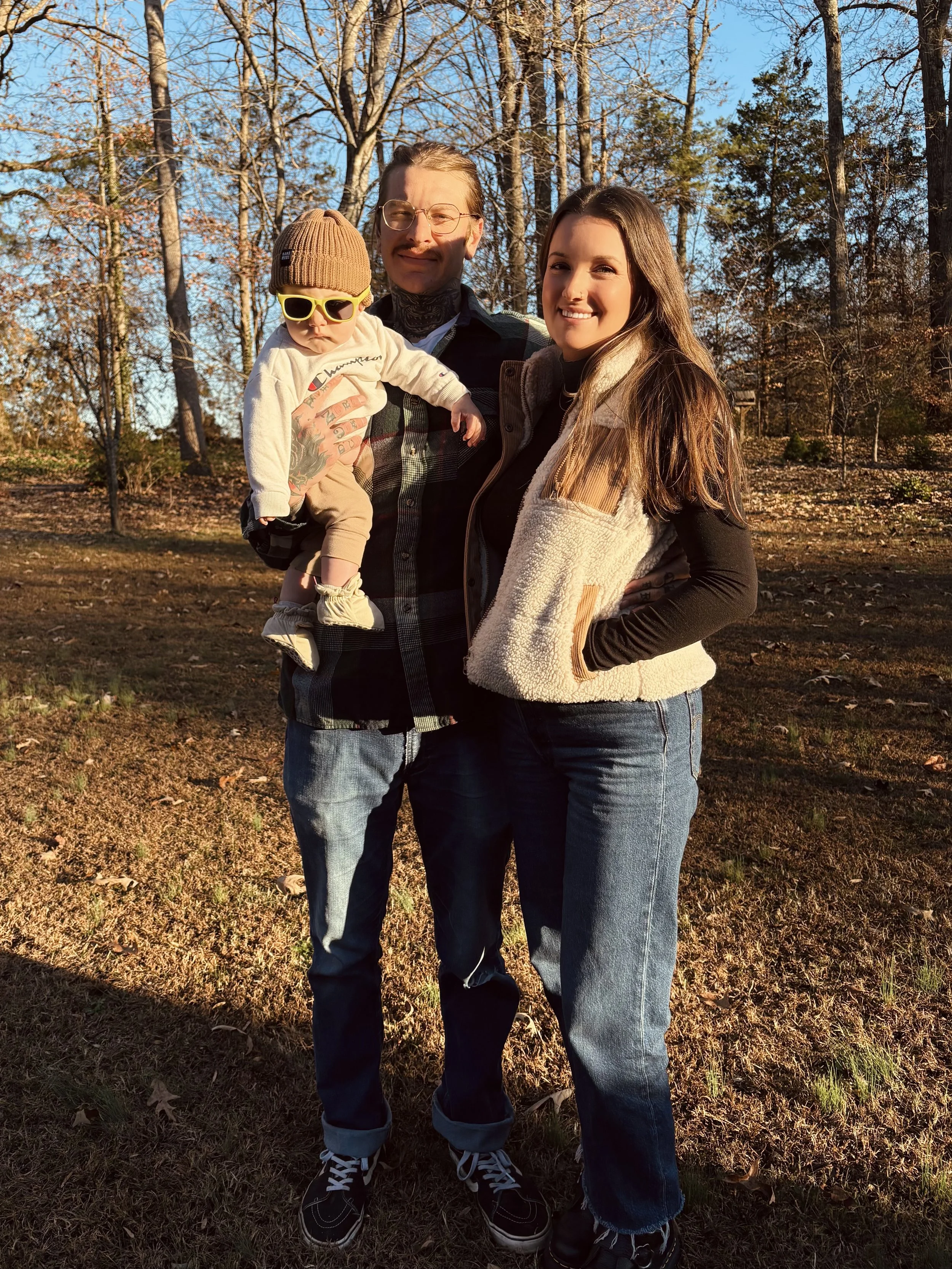 A family of three standing outdoors in a wooded area during autumn, with trees and fallen leaves in the background, enjoying a sunny day.