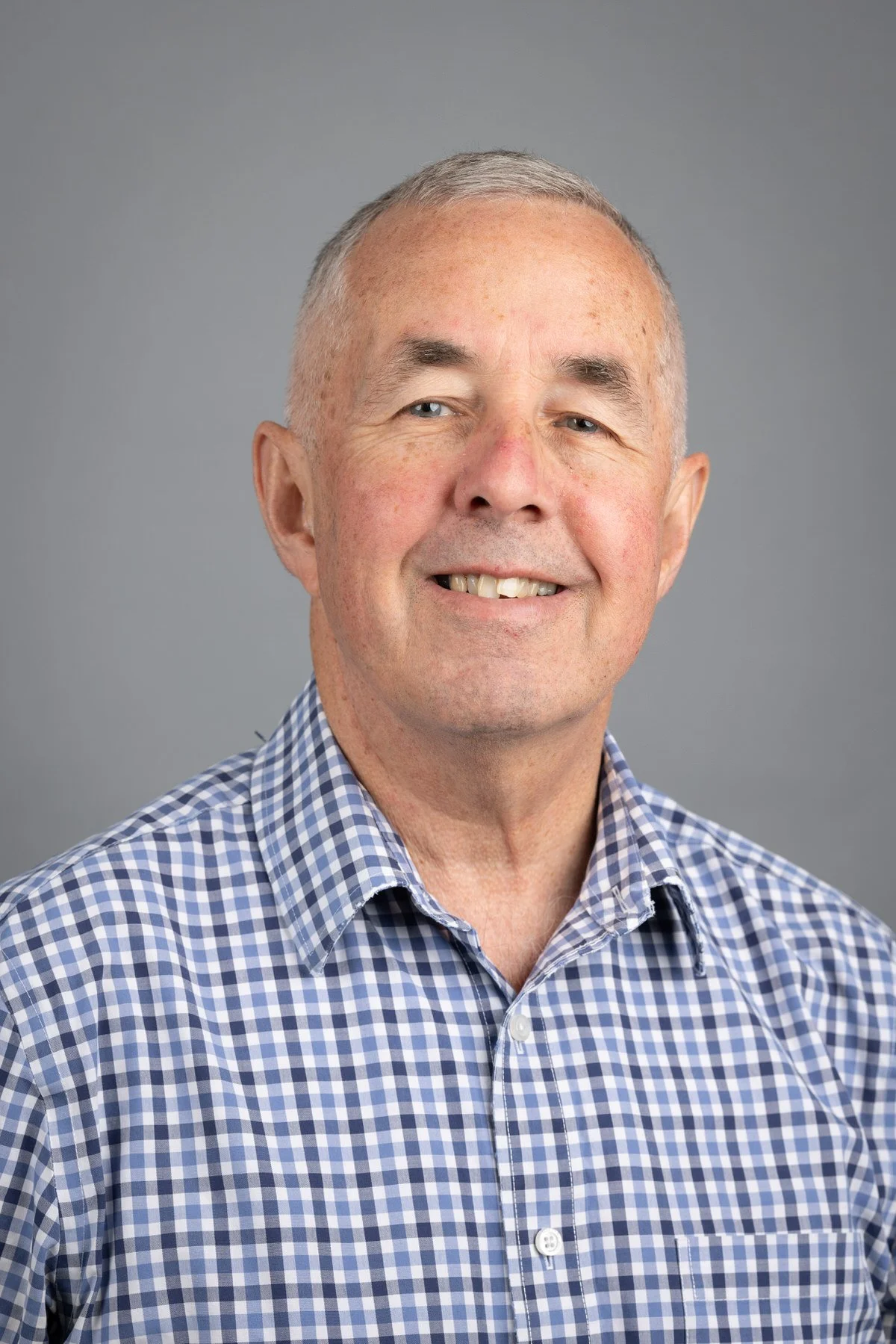 Portrait of an older man with glasses, wearing a plaid shirt and a patterned sweater, standing against a gray background.