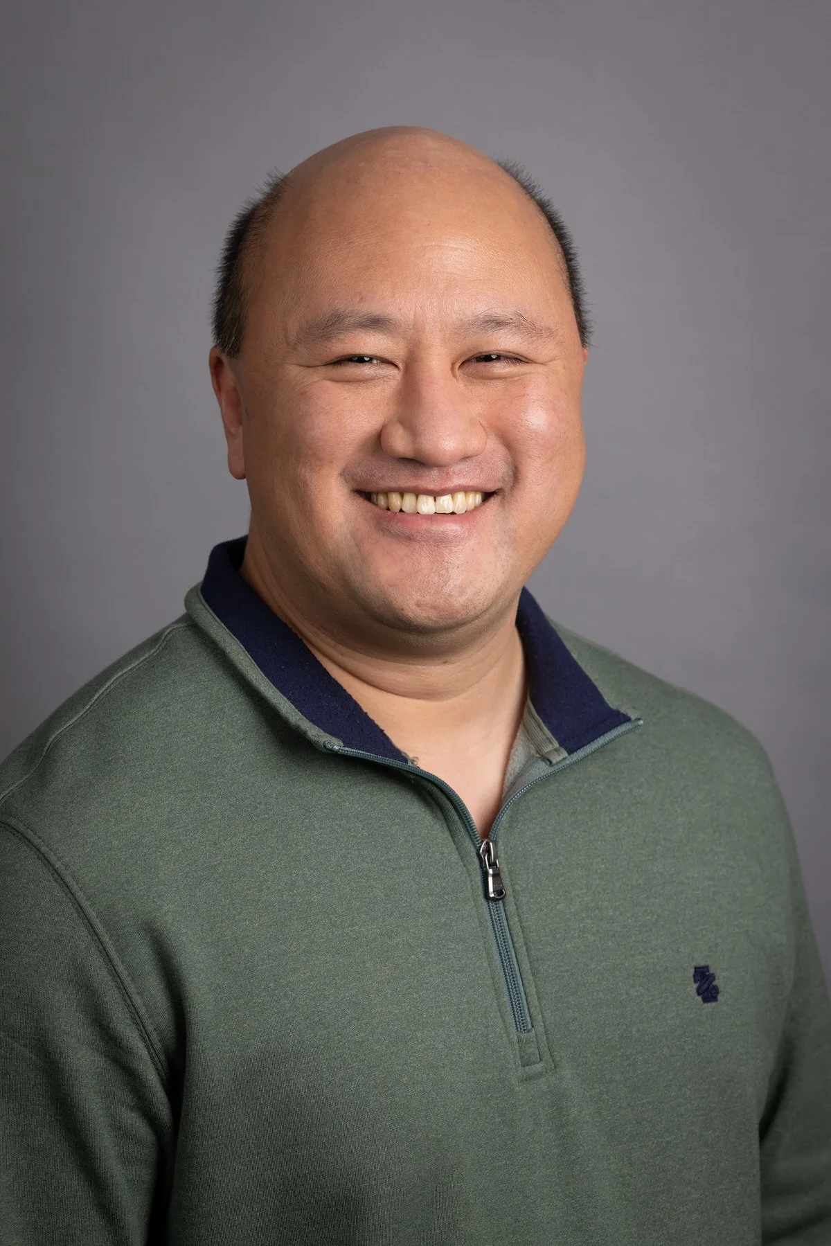 Headshot of man with dark hair, beard, and mustache, wearing a plaid shirt, smiling, against a plain gray background.