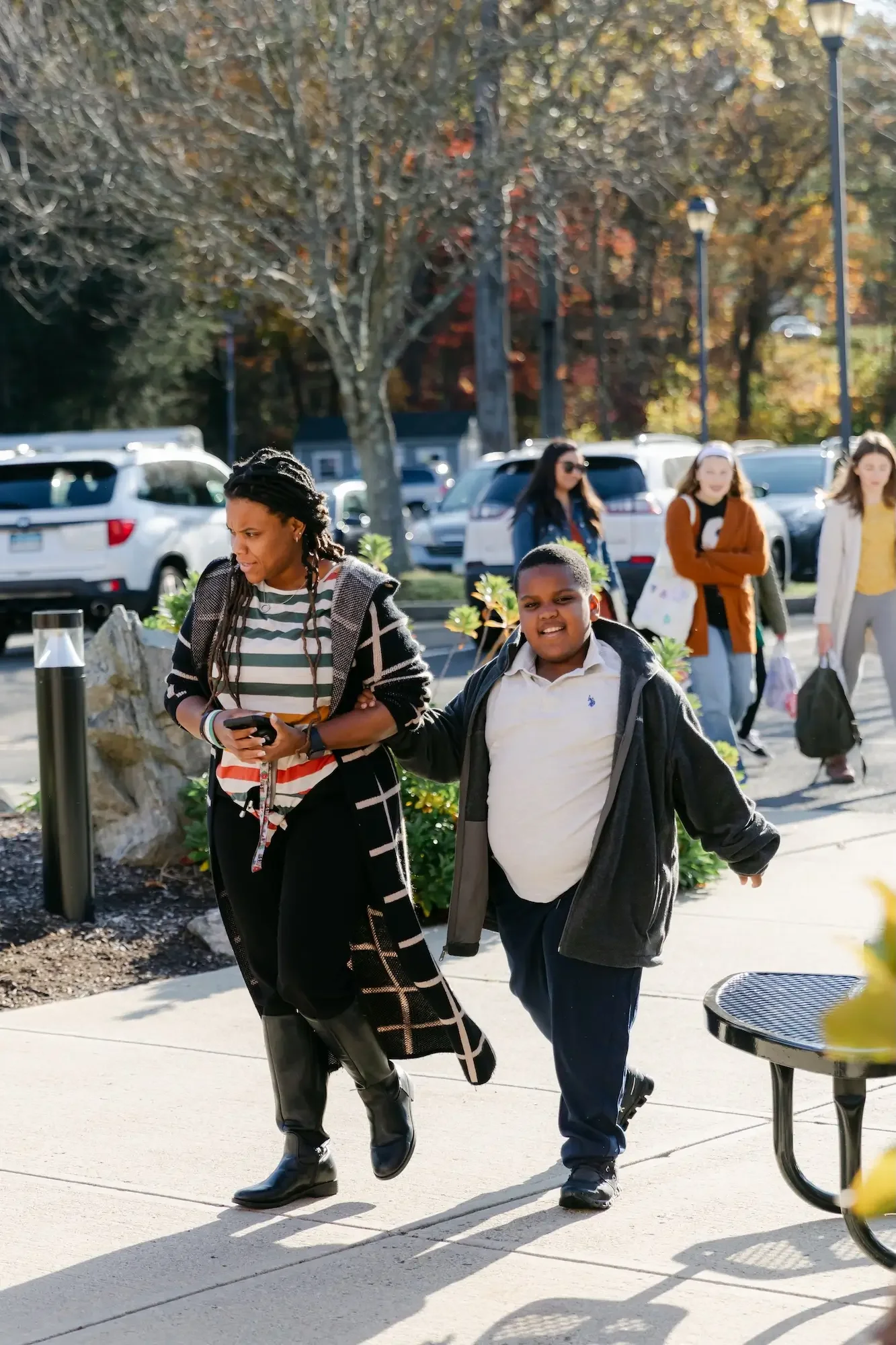 A woman walking with a young boy on a sidewalk in a parking lot with cars and other pedestrians in the background, during autumn.