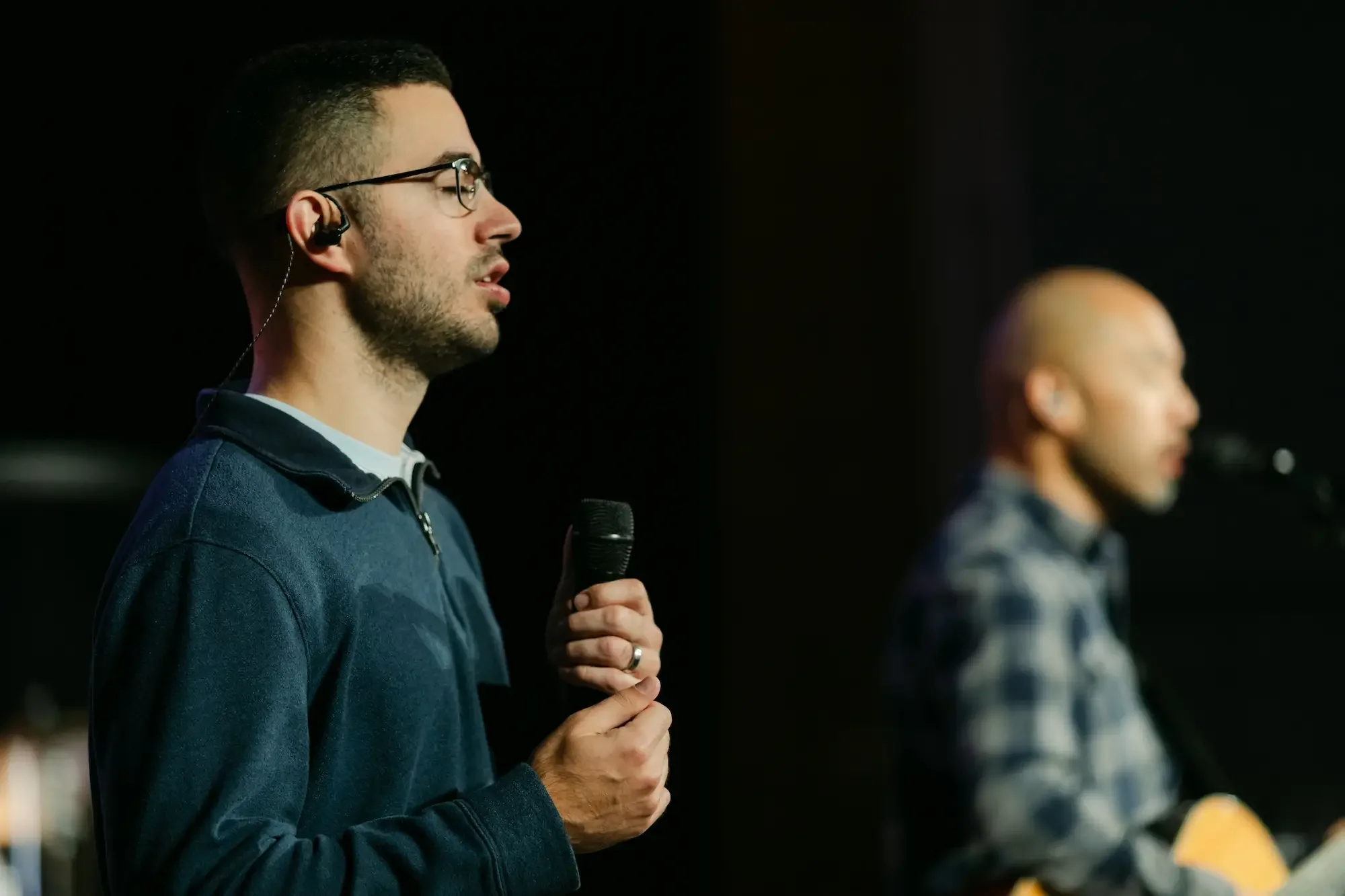 Two men with microphones, one in the foreground with glasses and headphones, and another in the background, singing or speaking, on a dark stage.