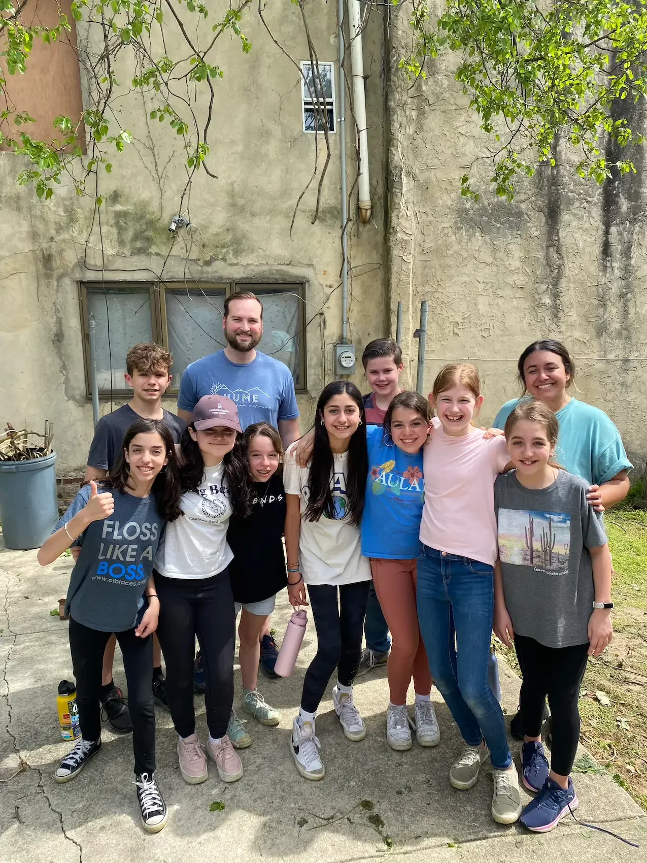 A group of young girls and two adults posing outdoors, smiling and standing close together in front of an old building with a concrete yard.