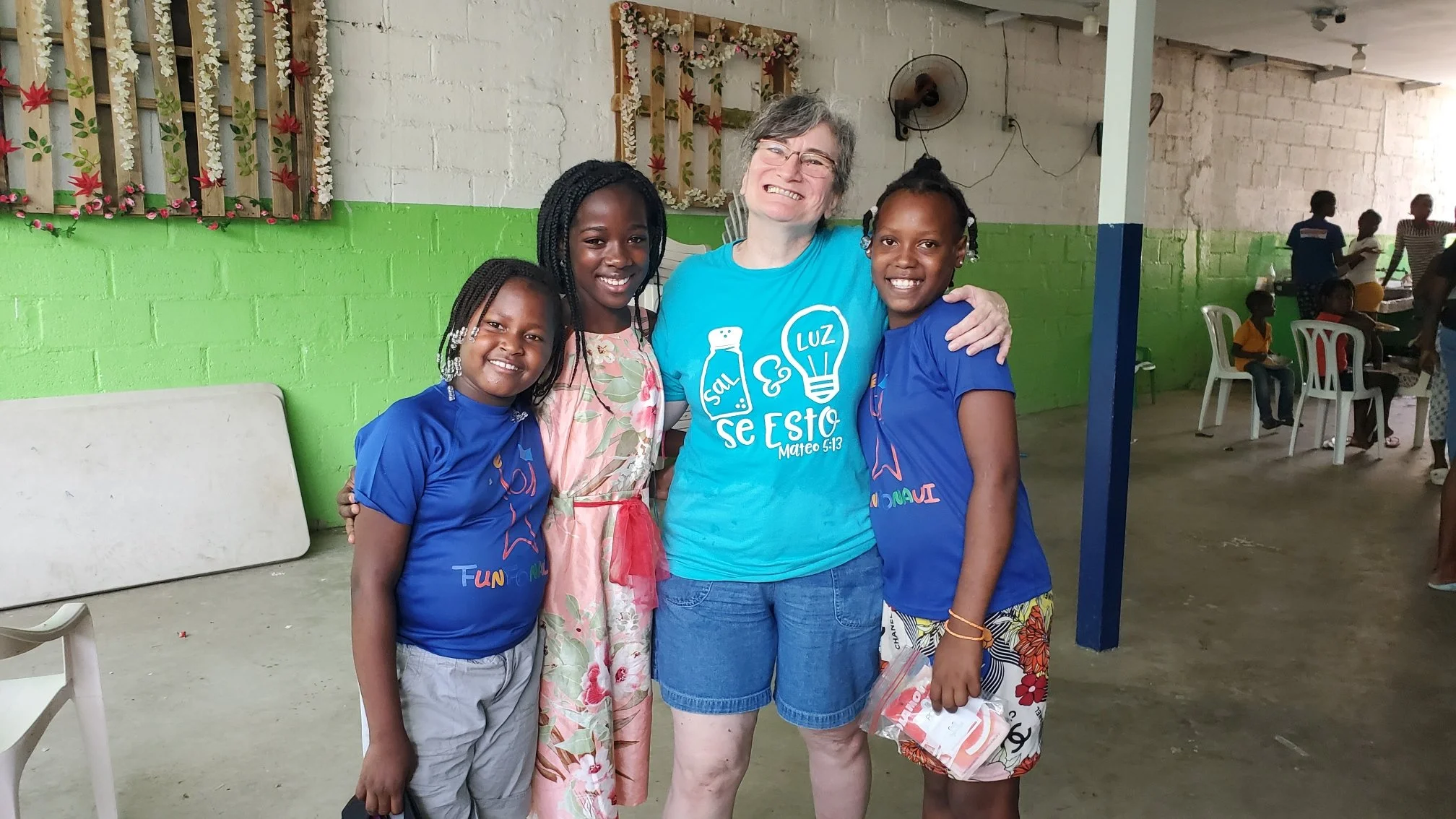 Four girls and one woman smiling and hugging in a room with green and white walls, some chairs, and other children in the background.